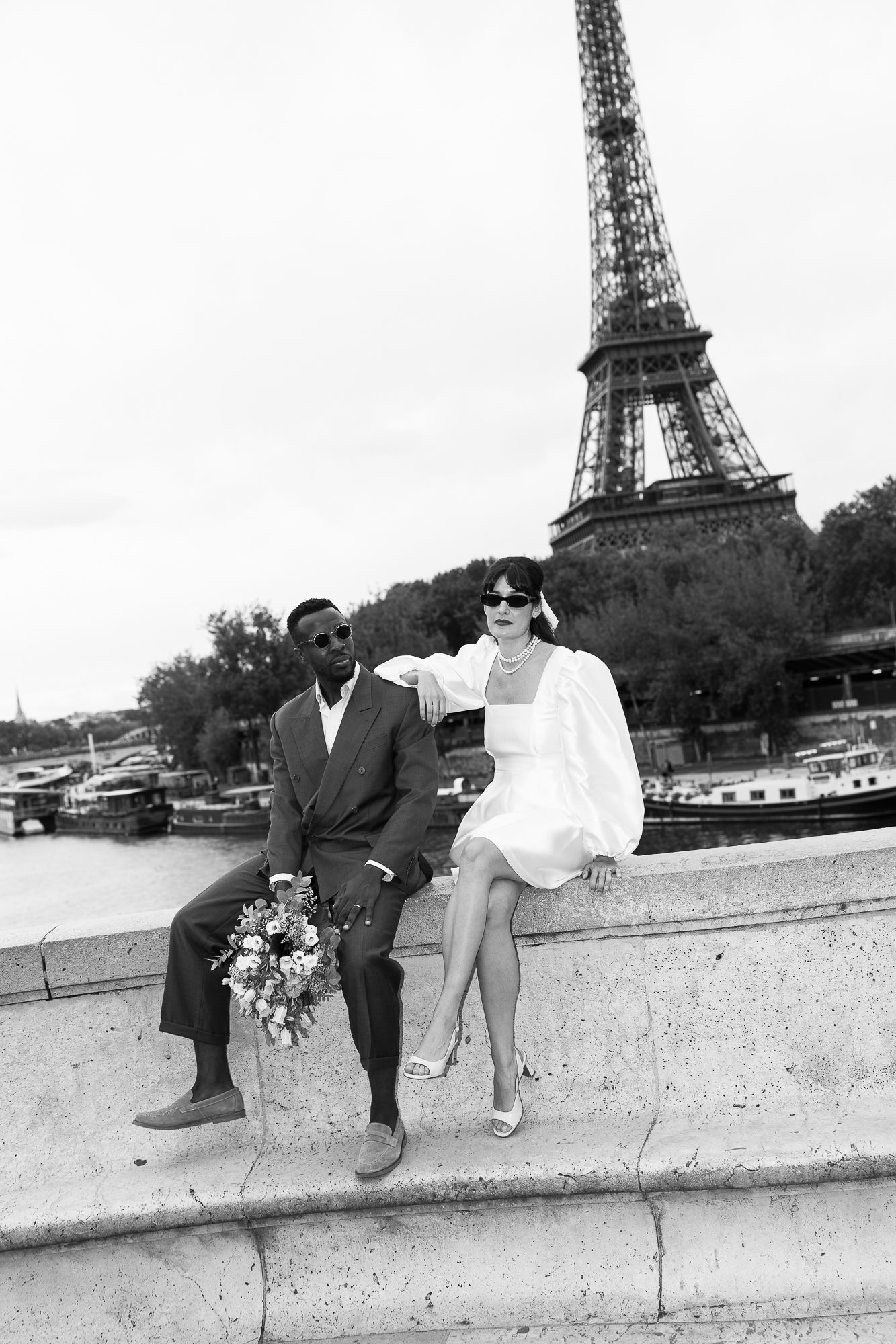 black and white photo shows a couple sitting on a wall with the Eiffel Tower visible in the background in Paris.