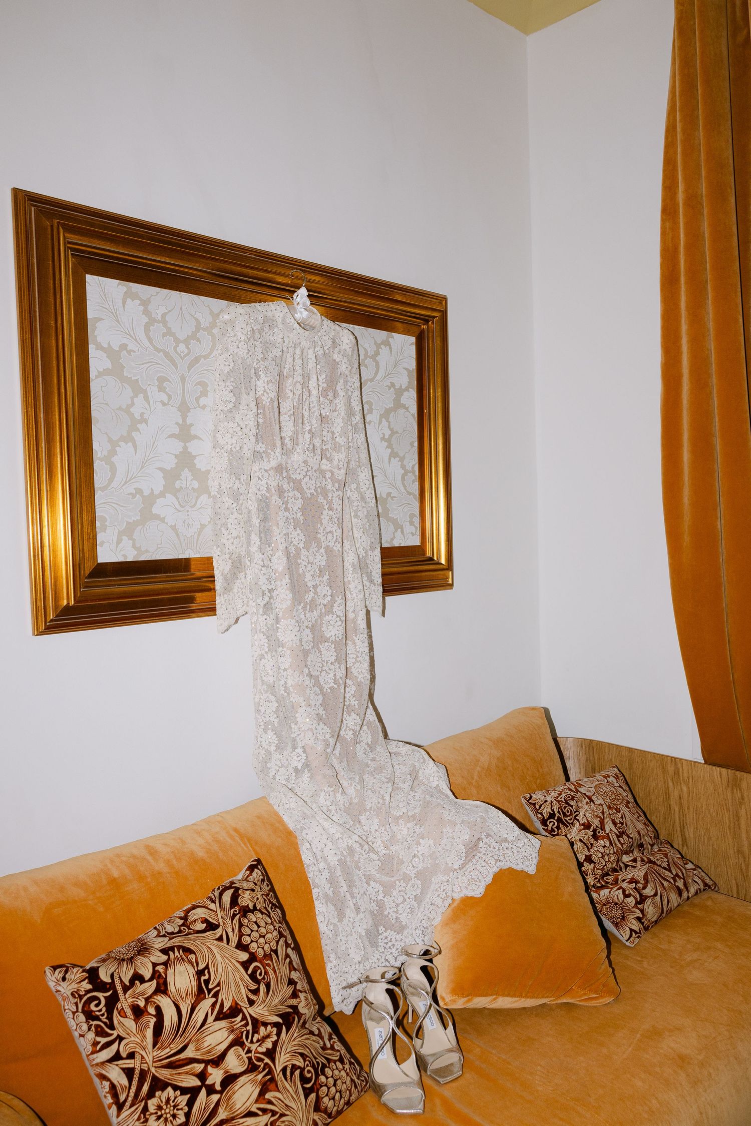 Delicate lace wedding dress hanging on gold-framed mirror above orange velvet couch.