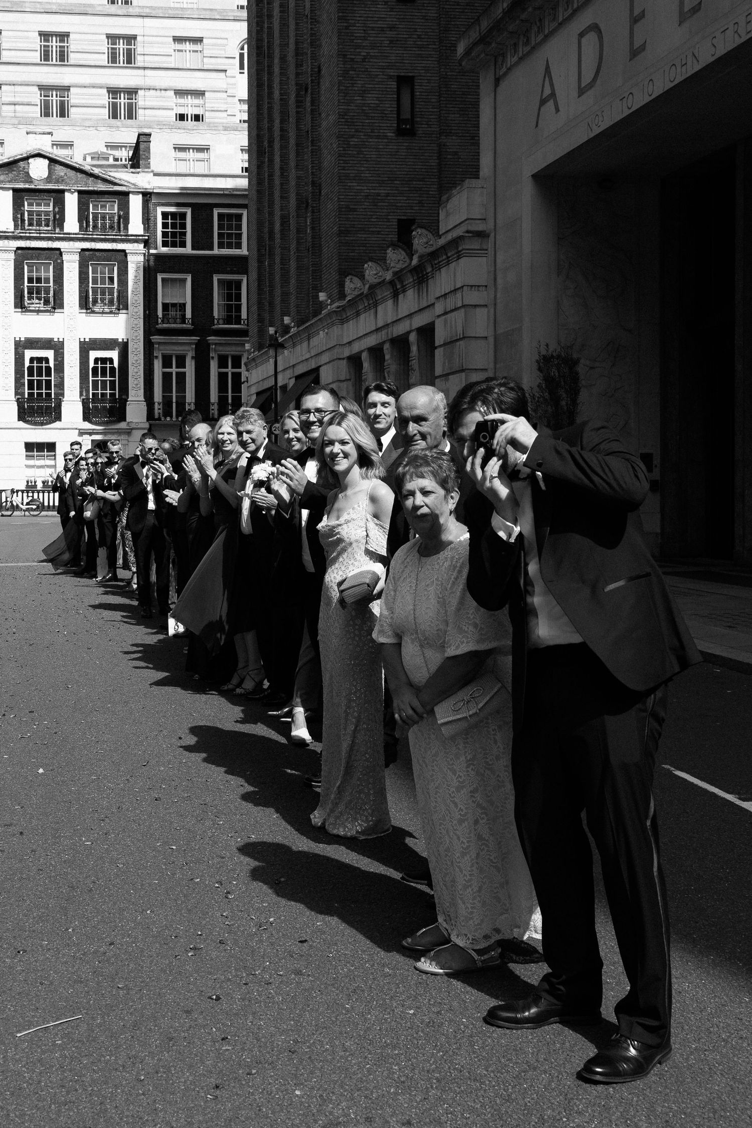 A black and white photograph of photographers lined up on a street near historic buildings raising their cameras.