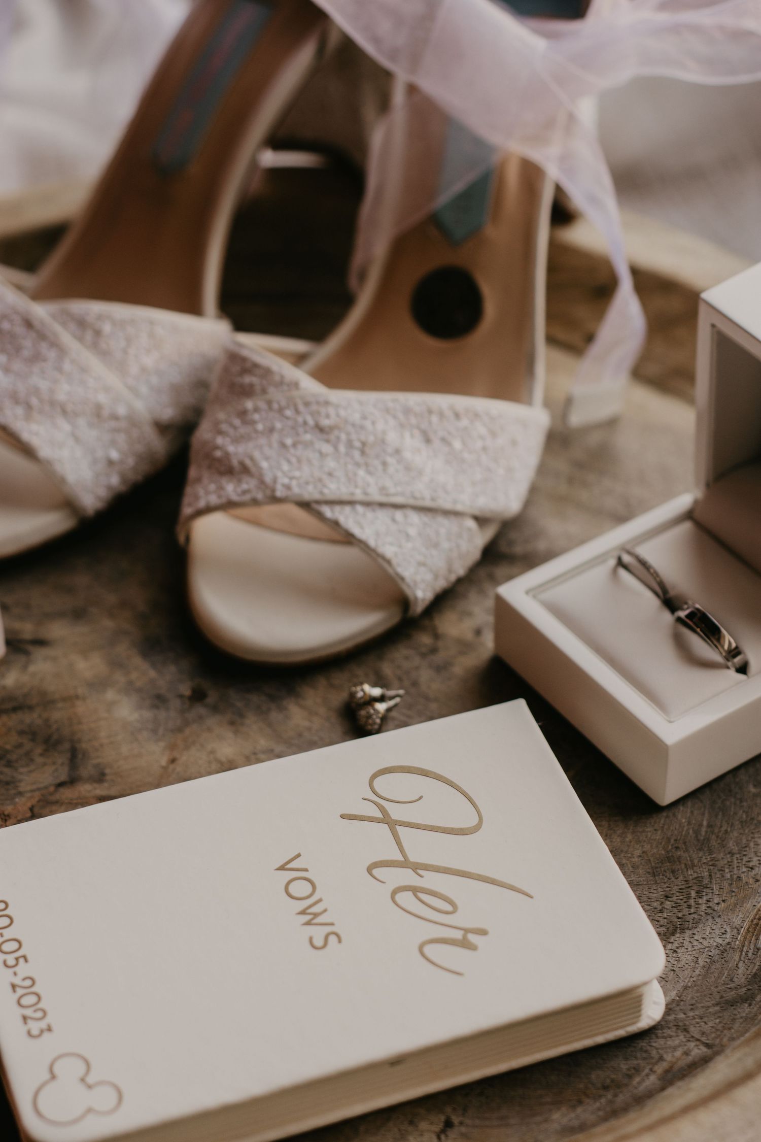 Wedding details including white lace shoes, rings and vows book arranged on rustic surface.