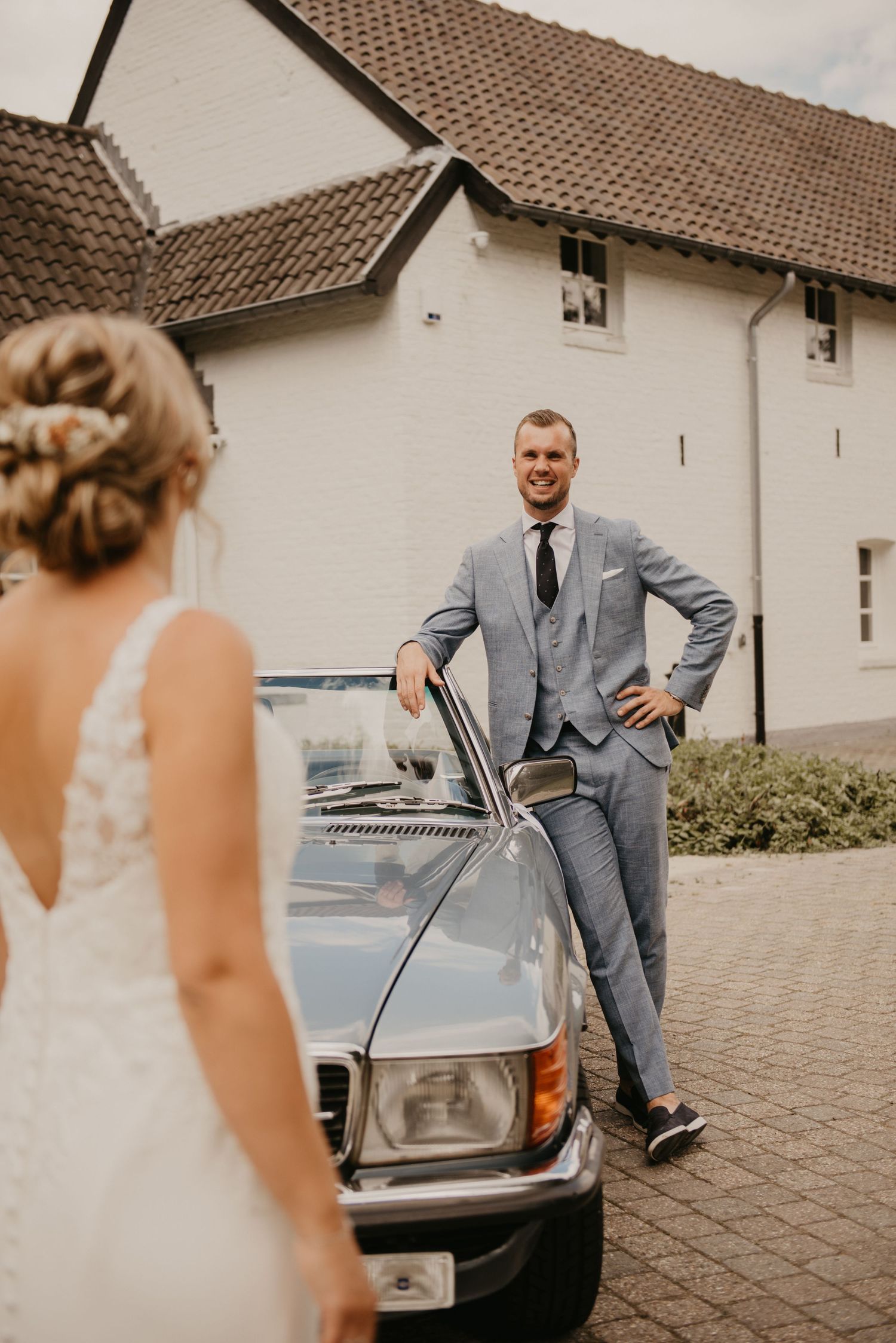 Wedding couple poses with a classic silver car outside a white building.