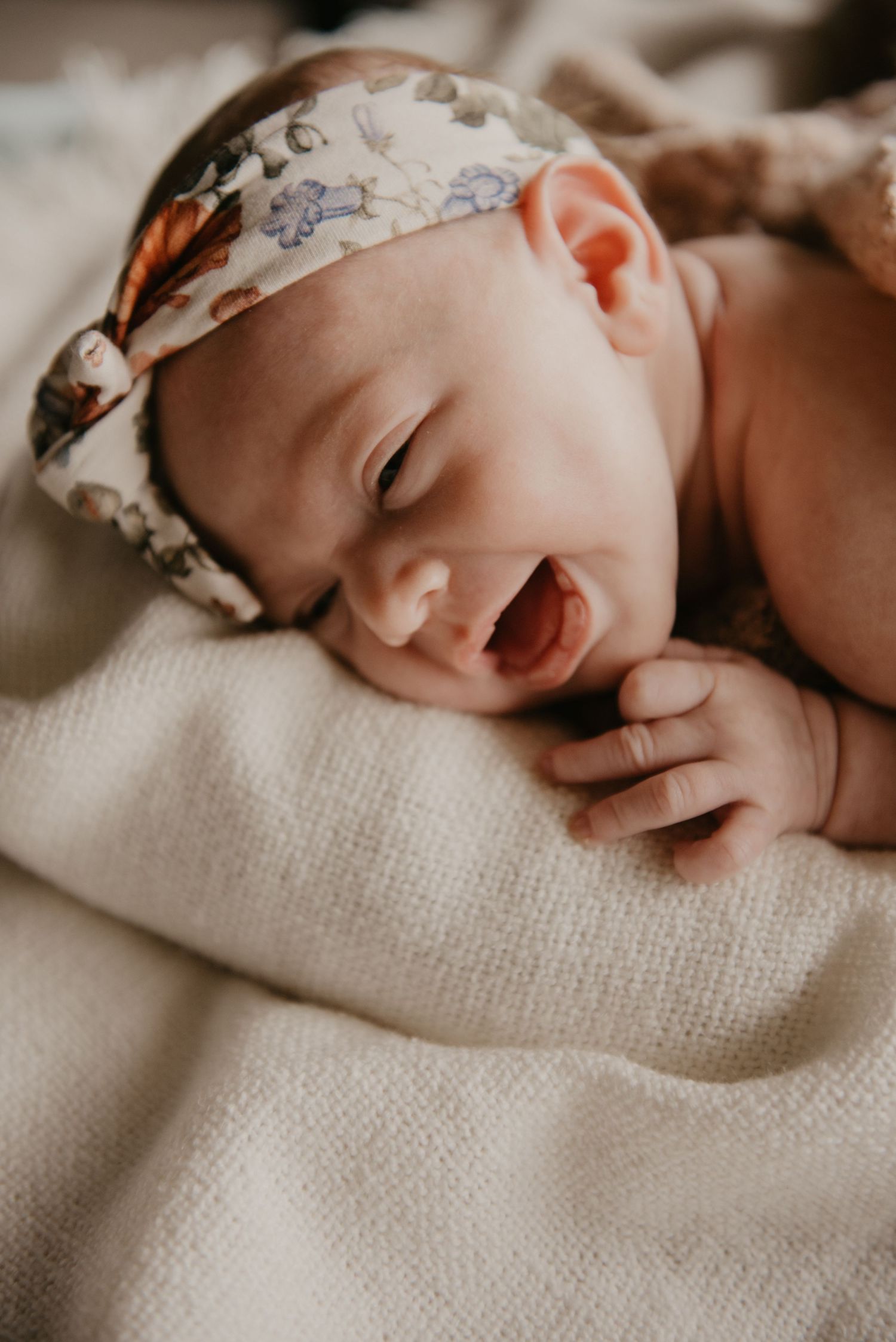 Newborn baby wearing floral headband lying peacefully on textured beige fabric.