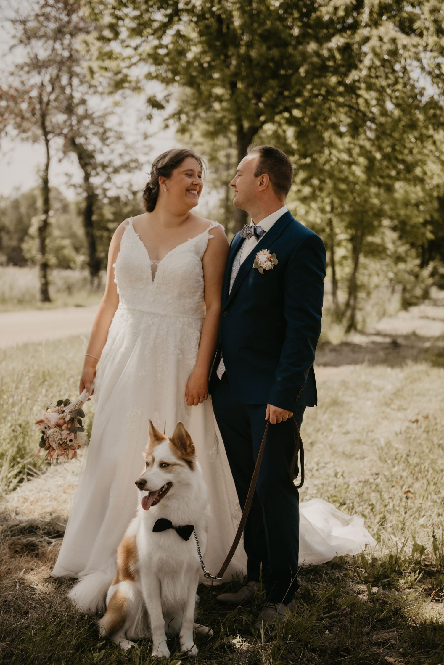 Wedding couple poses outdoors with their white dog in a sunny natural setting.