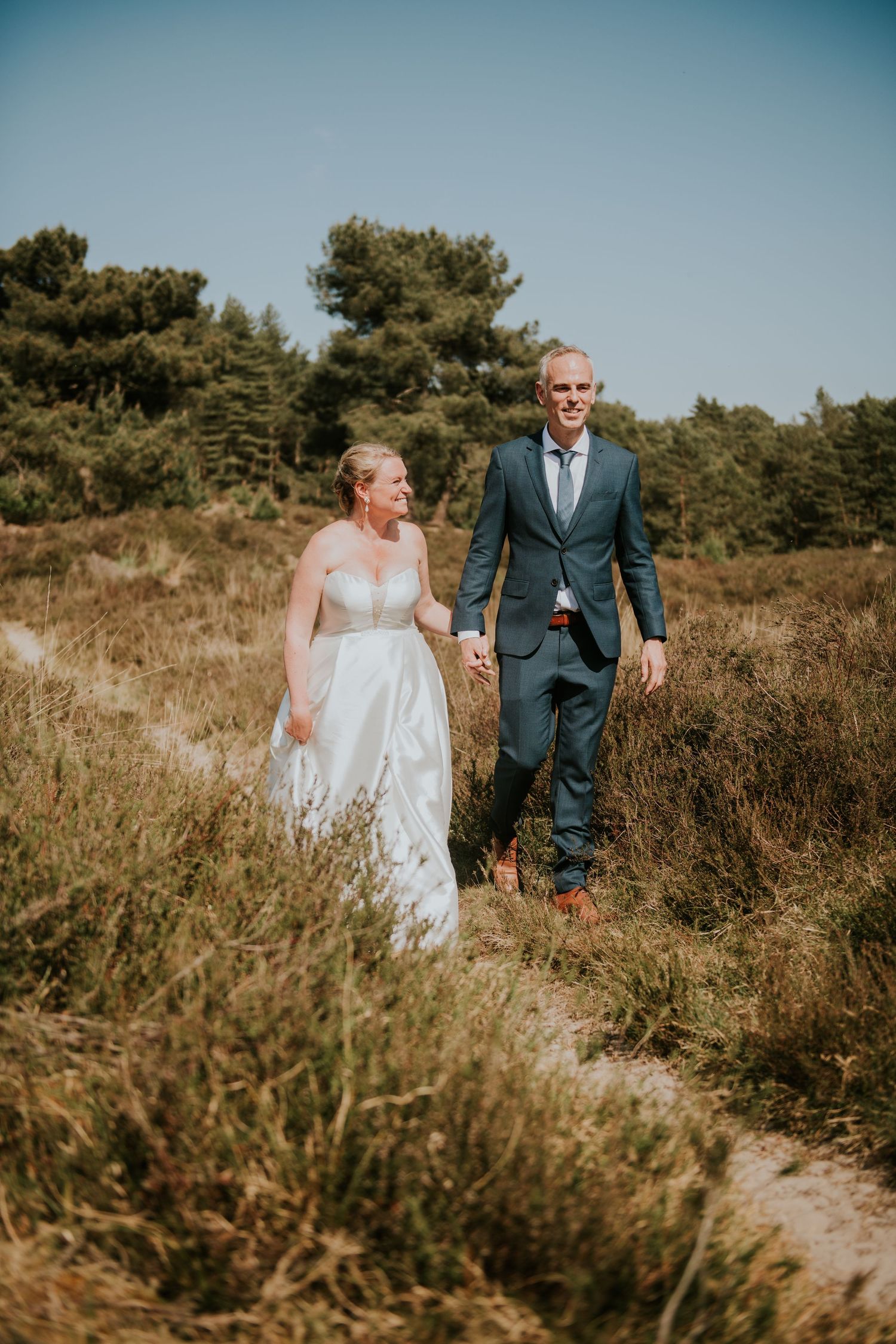 Couple walks hand in hand through tall grass on a scenic hillside path.