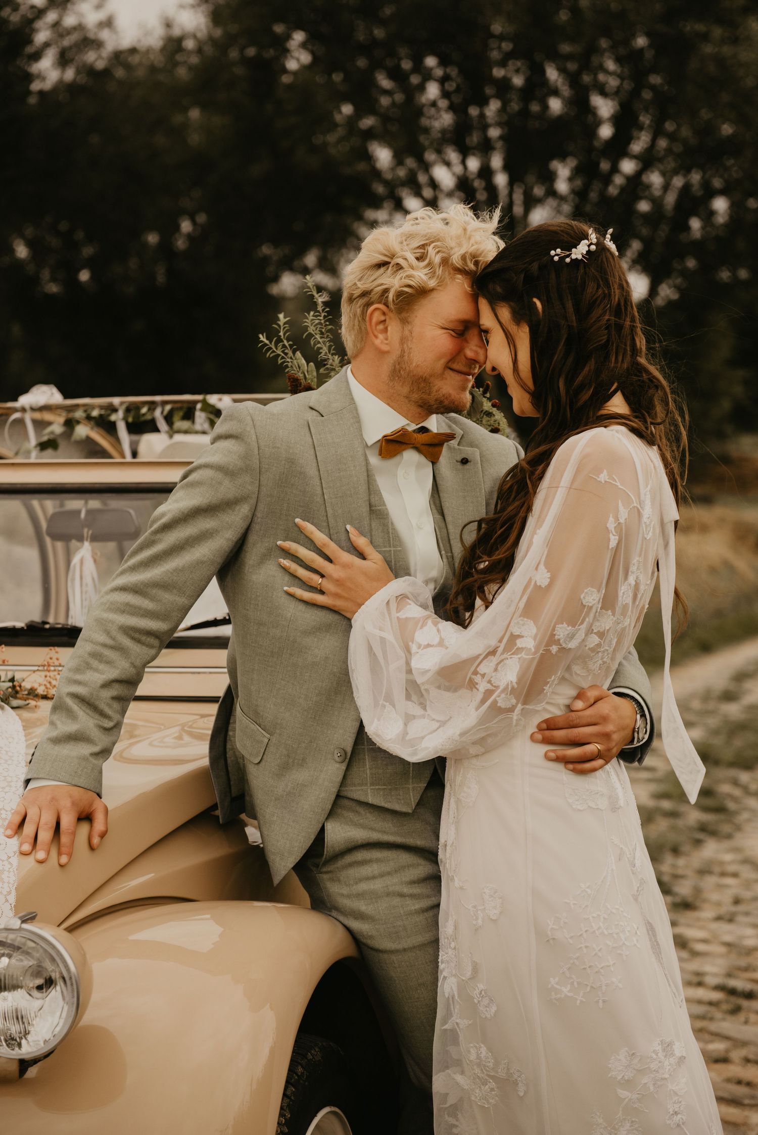 Couple in wedding attire poses against classic beige vintage car during sunset photoshoot.