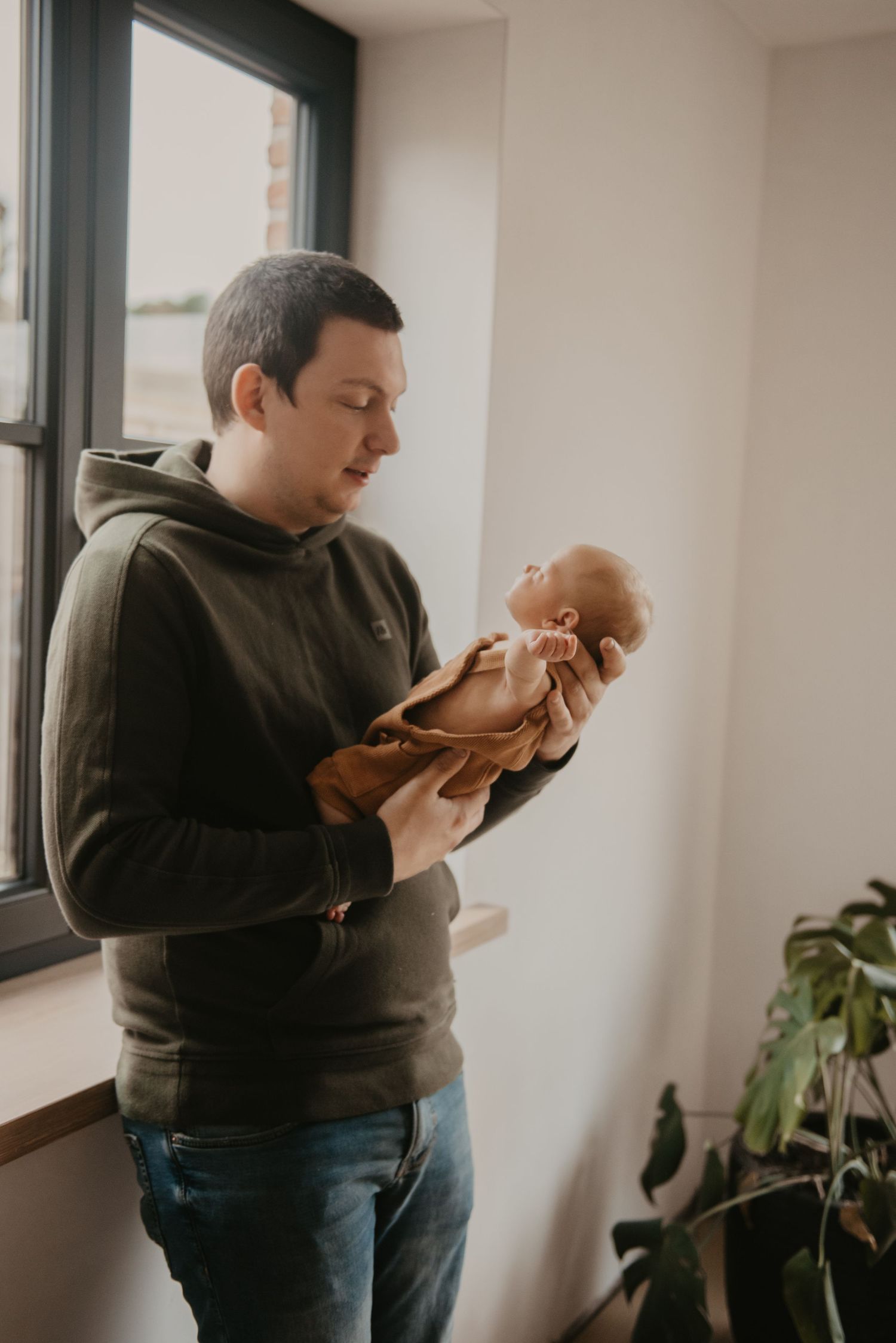 Parents sharing tender moment with newborn by window with houseplant.