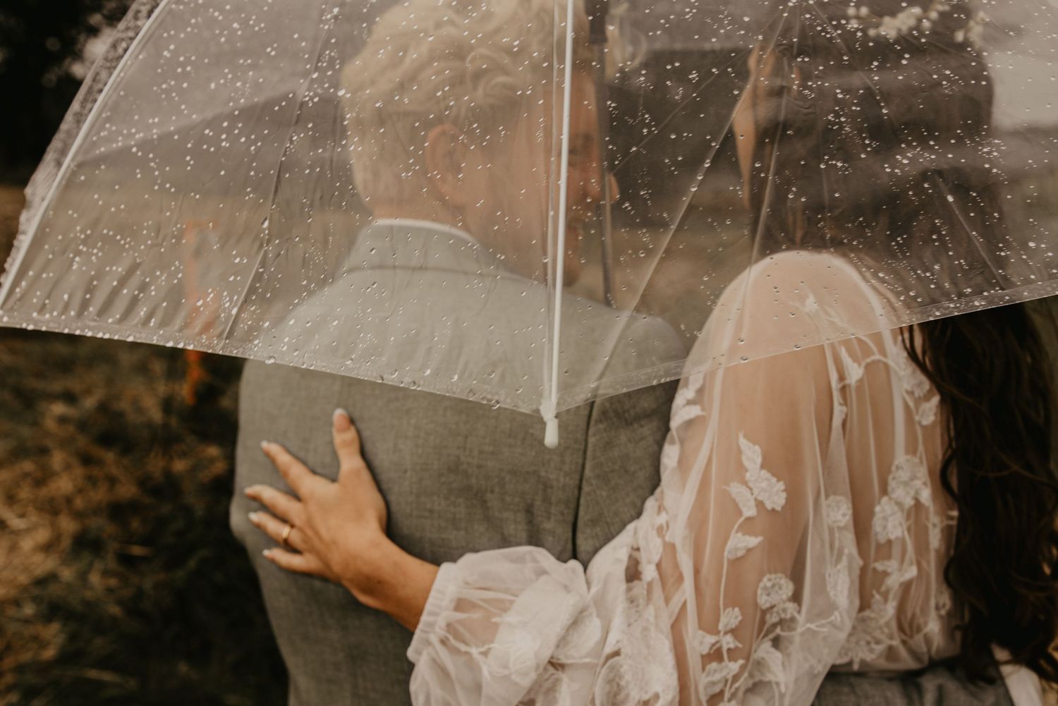 Romantic wedding couple shares intimate moment under clear umbrella in natural setting.