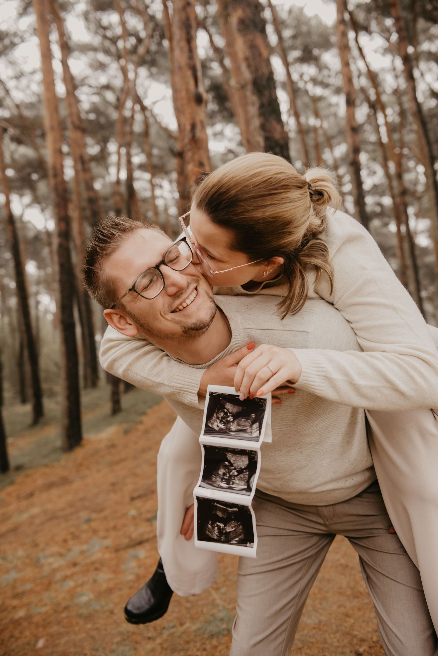 Playful couple in neutral sweaters sharing joyful moment with ultrasound photo in pine forest.