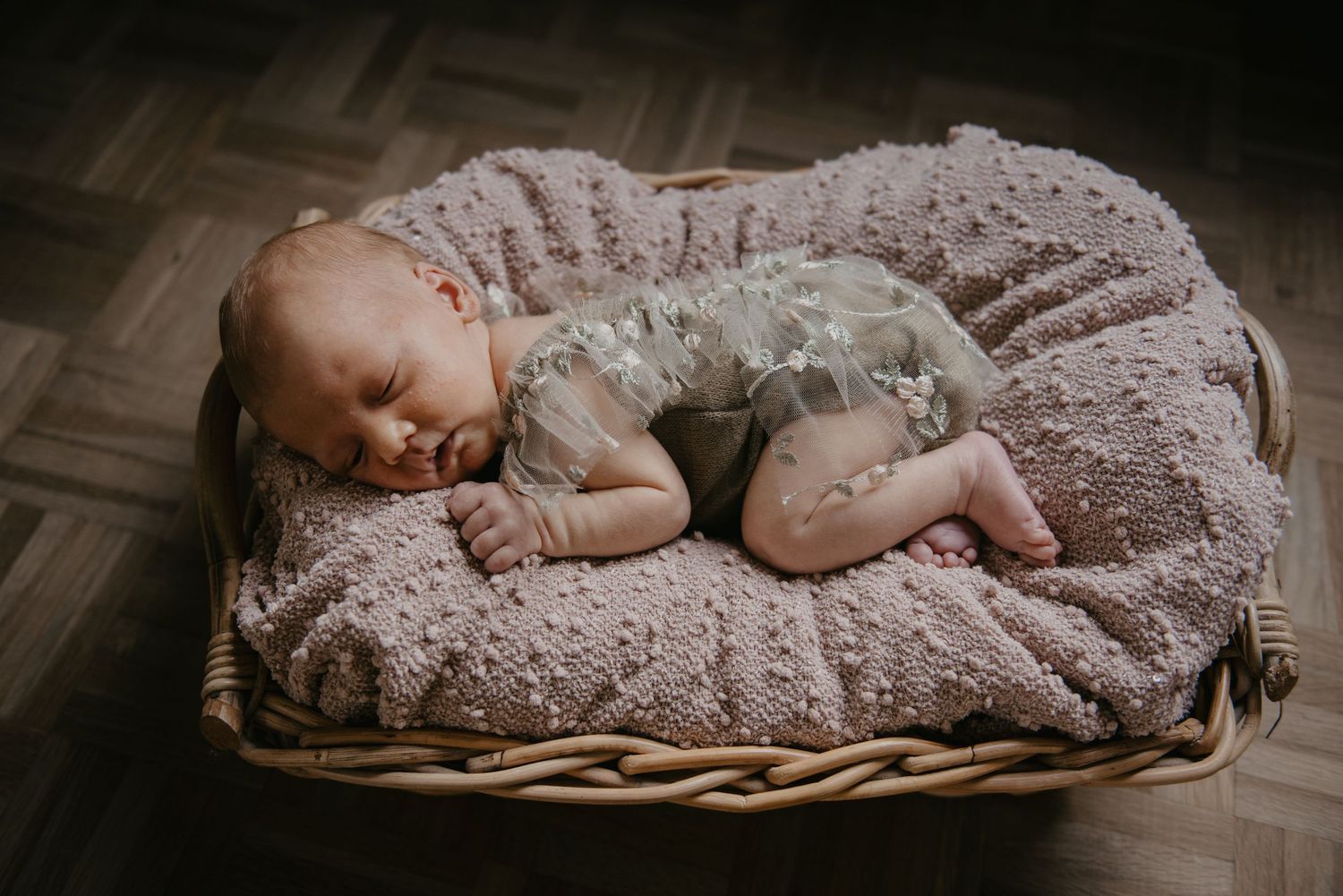 Sleeping infant in lace outfit resting on textured beige cushion in wicker basket.