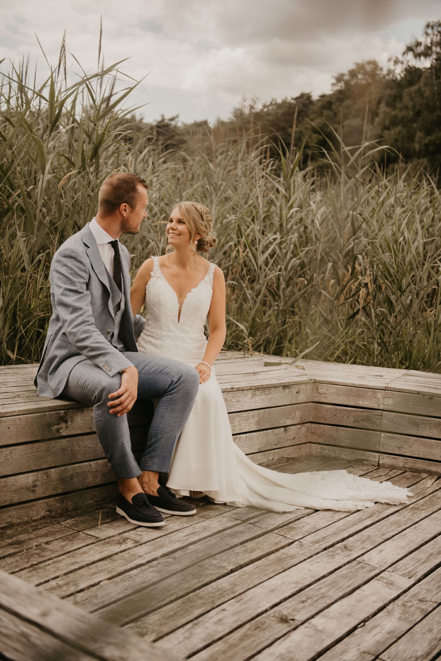 A couple sits together on a wooden deck surrounded by tall grass and reeds.