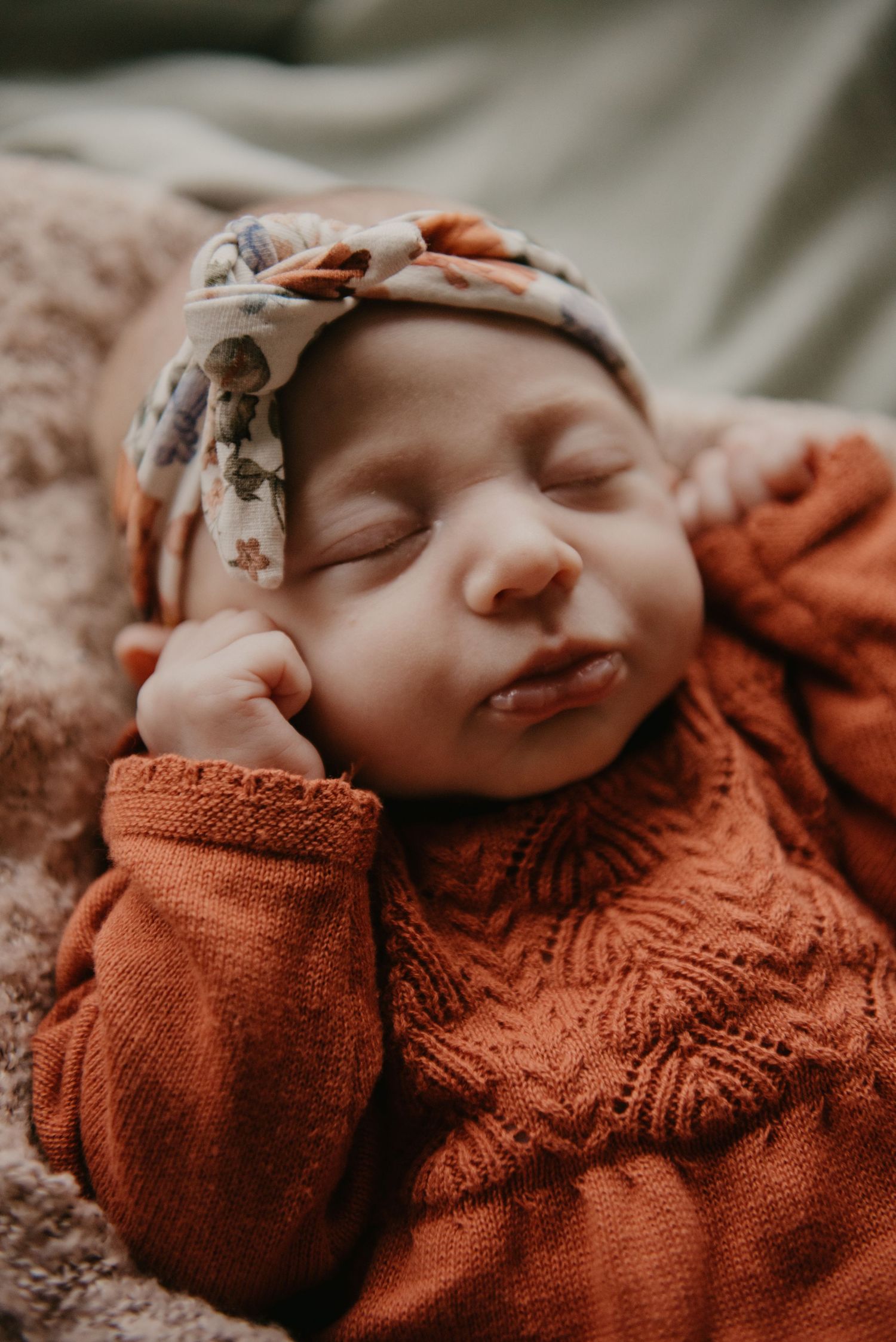 Sleeping baby wearing rust-colored knit sweater and floral headband on textured surface.