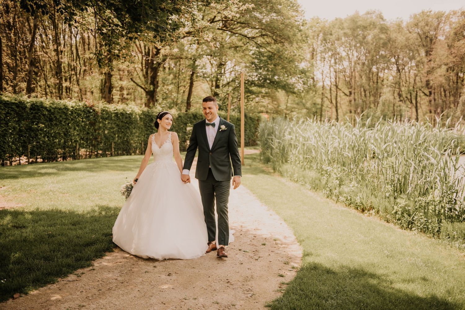 Bride and groom walking hand in hand on garden path surrounded by trees.