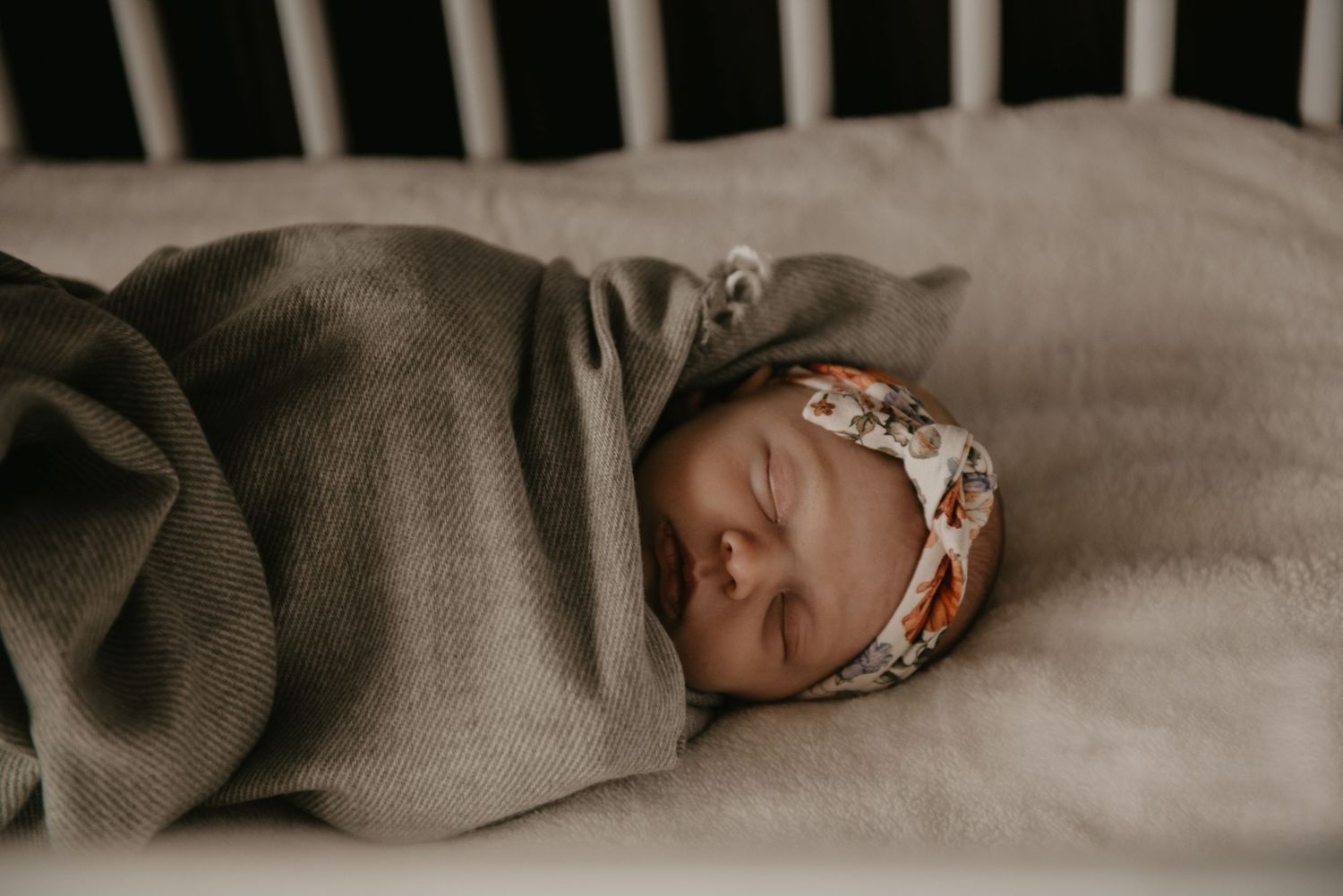 Peaceful newborn resting in crib with soft gray bedding and floral headband.