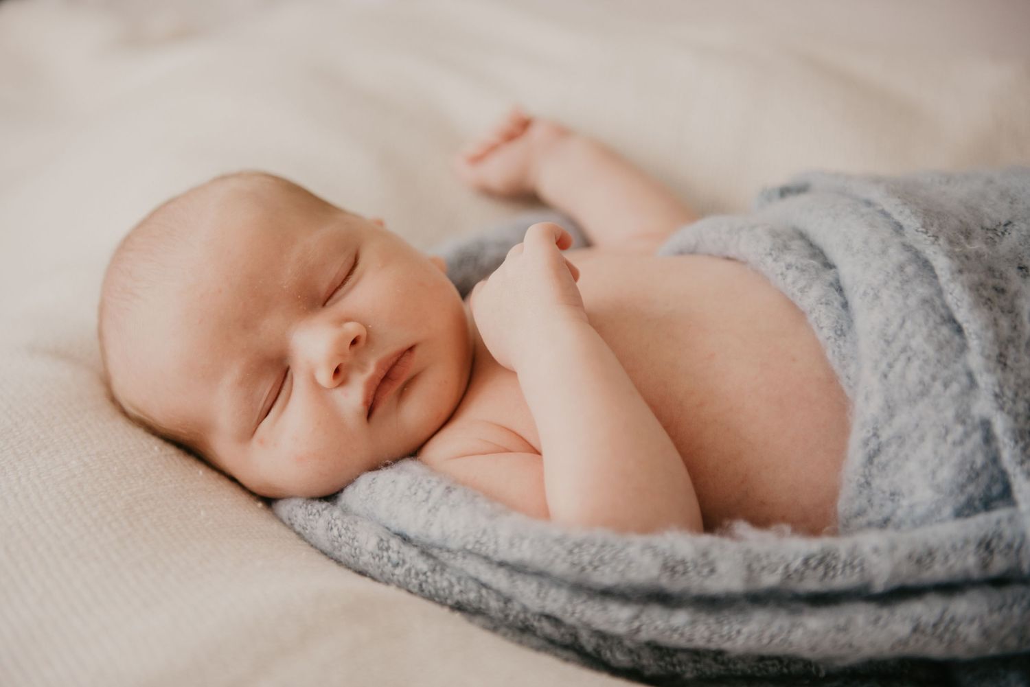 Sleeping baby wrapped in soft gray blanket on cream colored surface.
