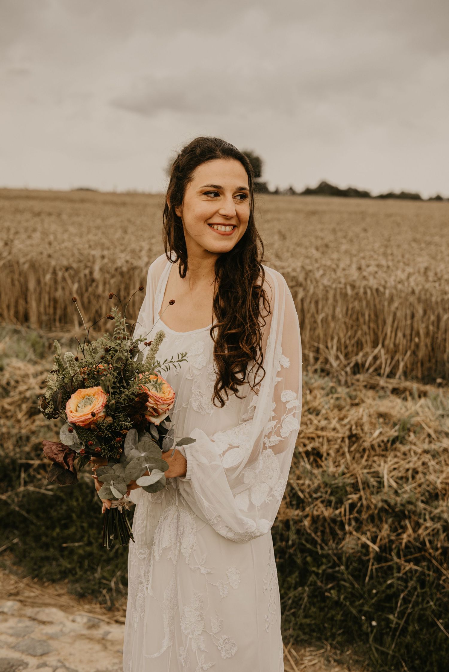 Bride in lace dress holds floral bouquet while standing in golden wheat field.