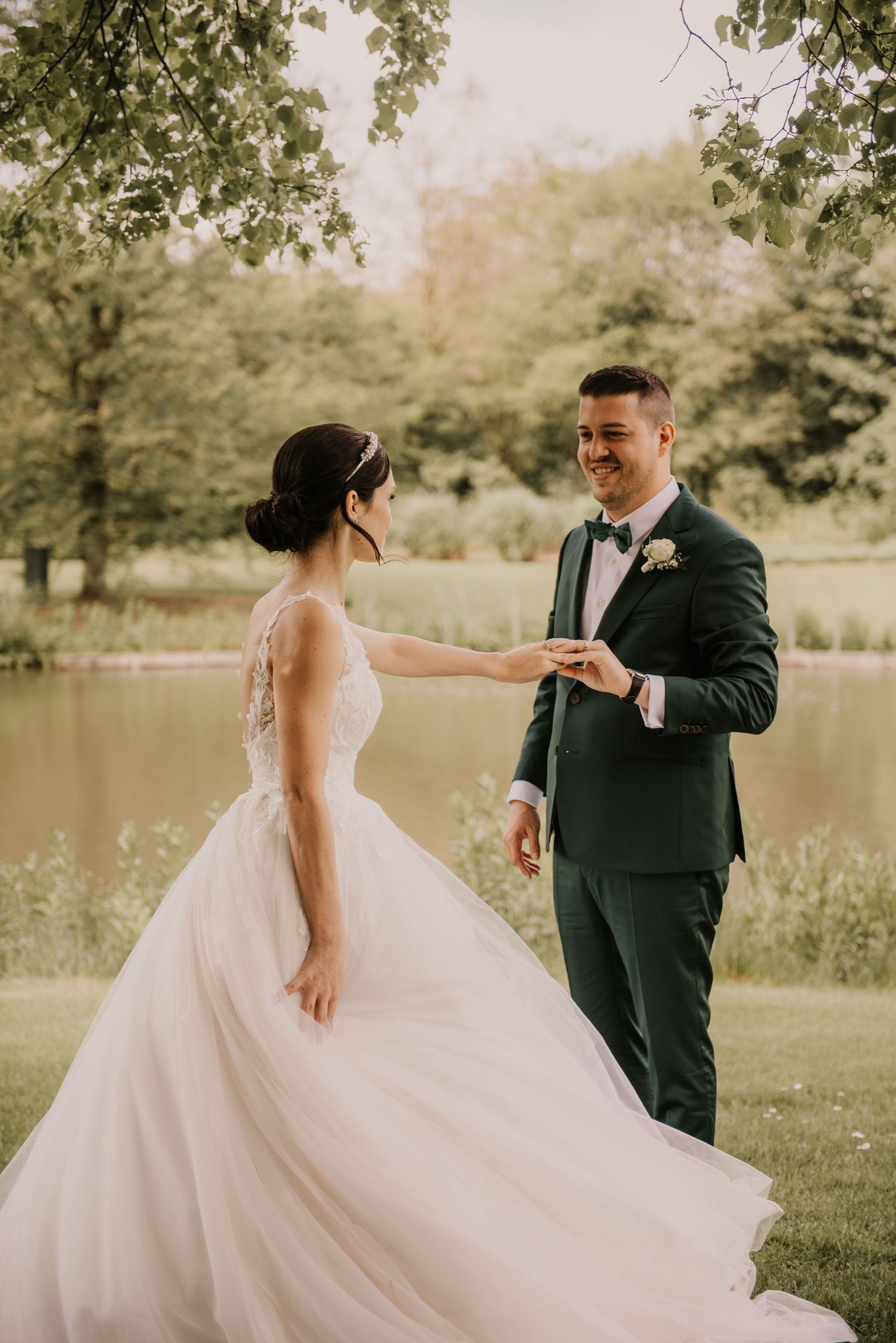 Bride and groom share romantic moment by lake in sunlit garden setting.
