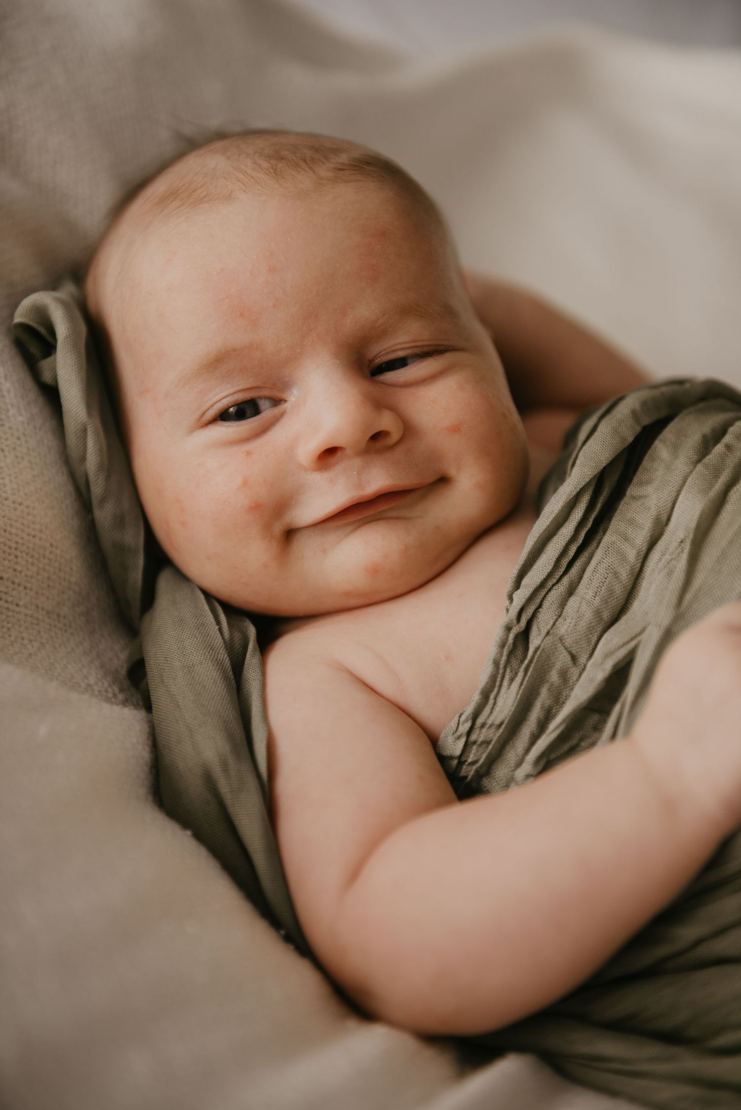 Baby wrapped in olive green fabric smiling while resting on soft bedding.