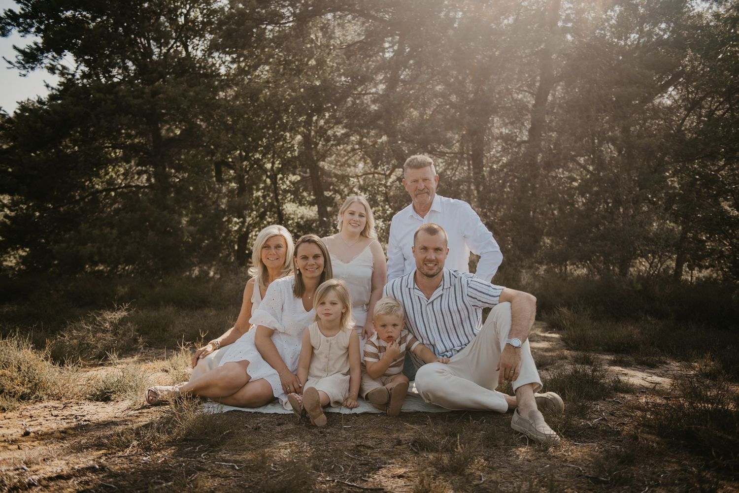 Family gathered together on ground during outdoor golden hour photo session in wooded setting.