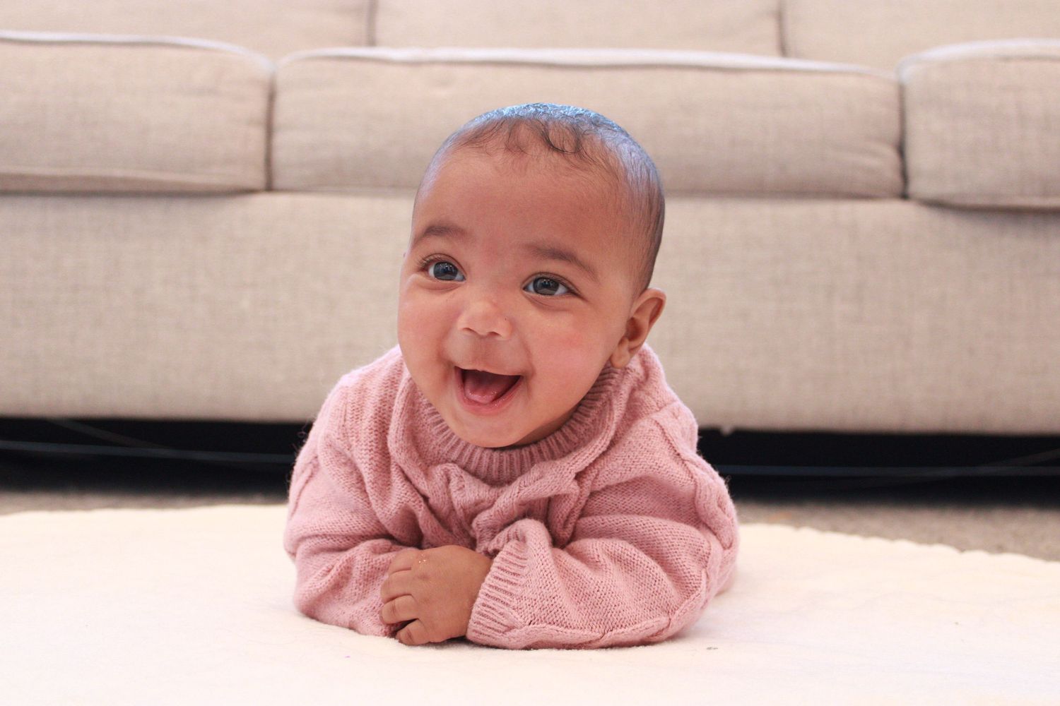 Baby in pink sweater smiling while lying on carpet.