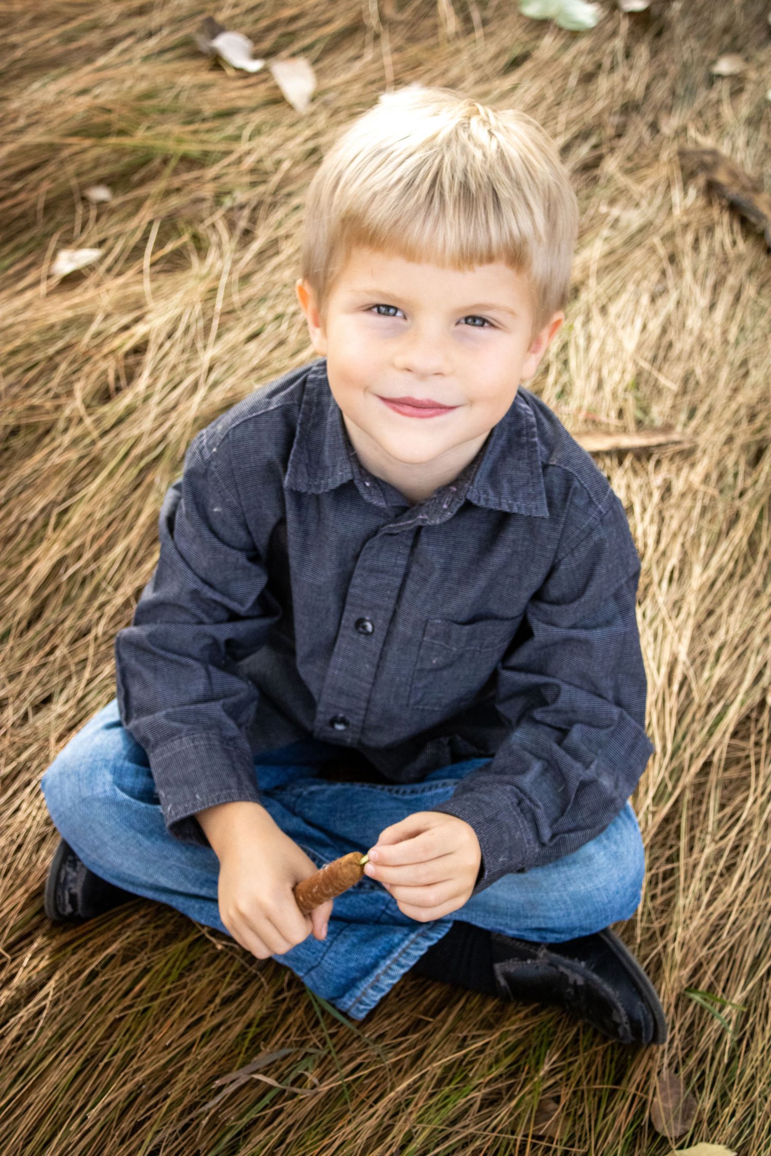A child wearing a navy button-up shirt and jeans sits in tall dried grass during an outdoor photo session.