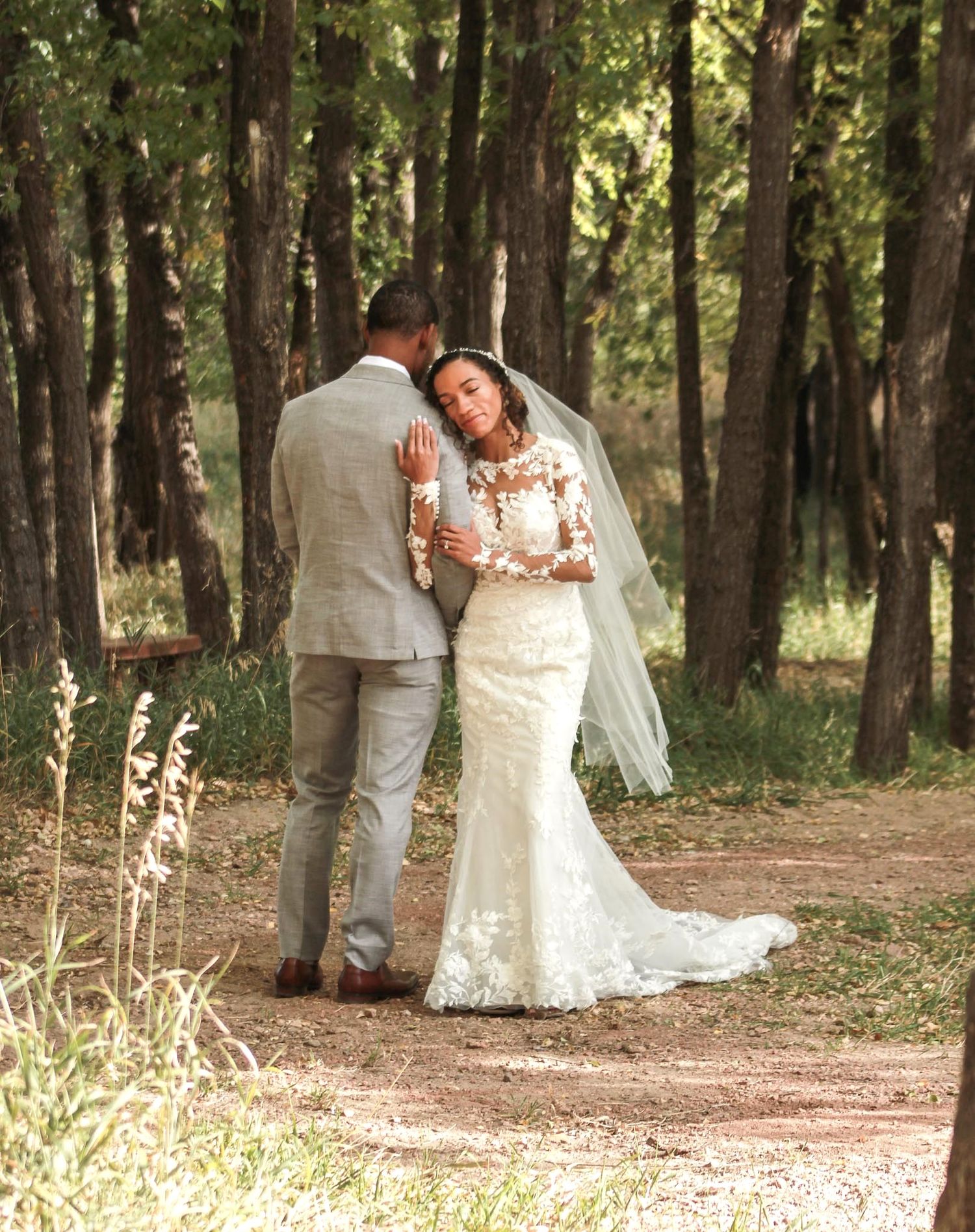 Wedding couple poses in a scenic wooded area with sunlight filtering through the trees.
