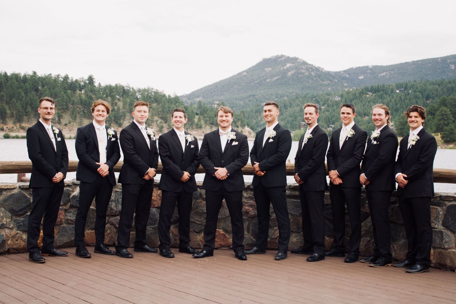 Groomsmen in black suits line up for a fun group photo on wooden dock by the river.