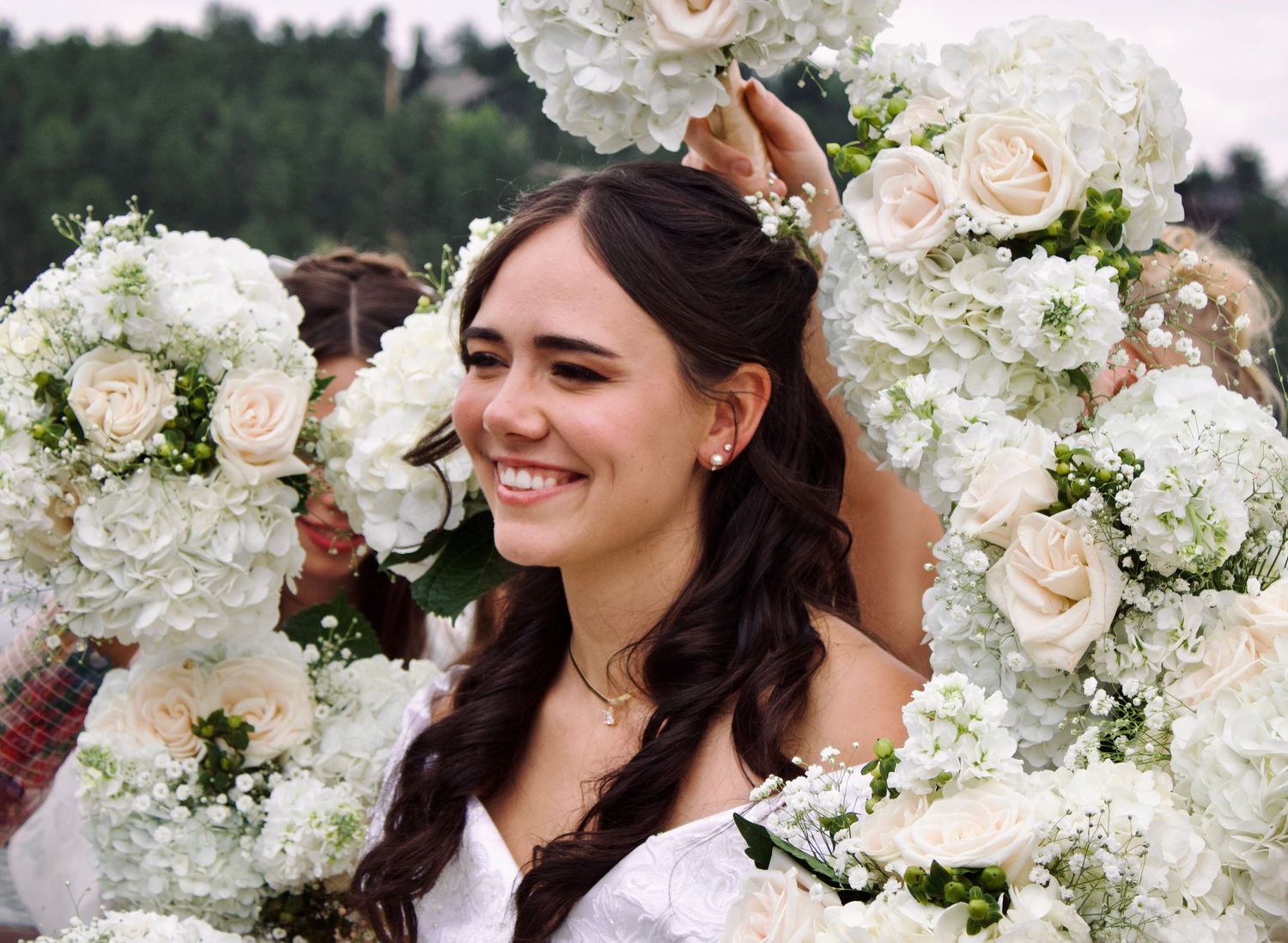 Elegant bride in white dress surrounded by archway of white roses and hydrangeas smiles brightly during wedding celebration.