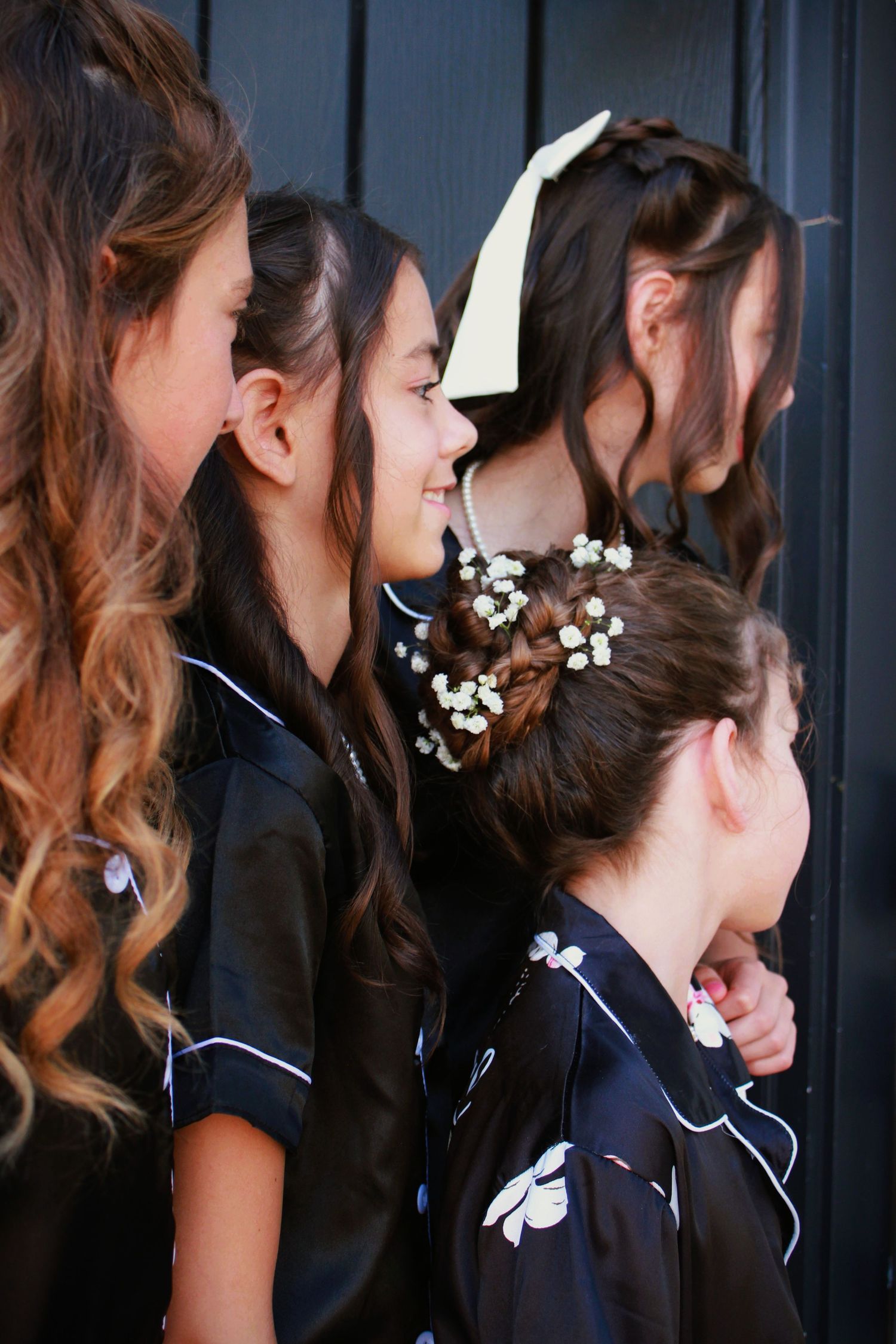 Group wearing matching dark robes with delicate white flowers woven into elegant hairstyles in profile view.