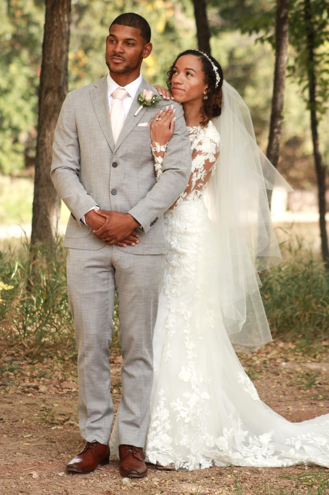 Wedding couple poses among trees in elegant grey suit and lace white gown.