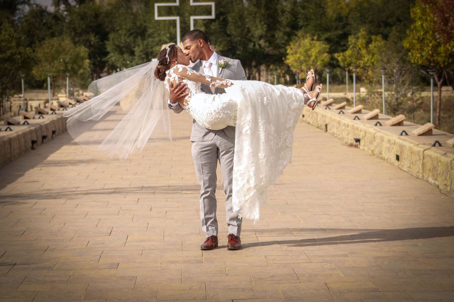 Wedding couple walks hand in hand on stone path with cross landmark in background.