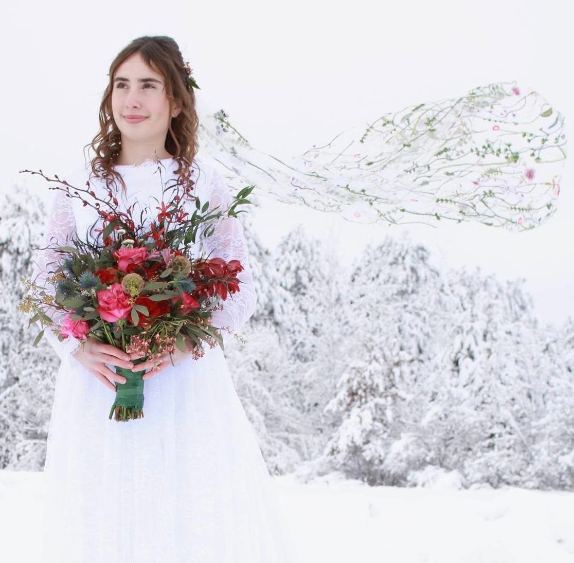 Winter bride in flowing white dress holds vibrant red bouquet against snowy mountain backdrop.
