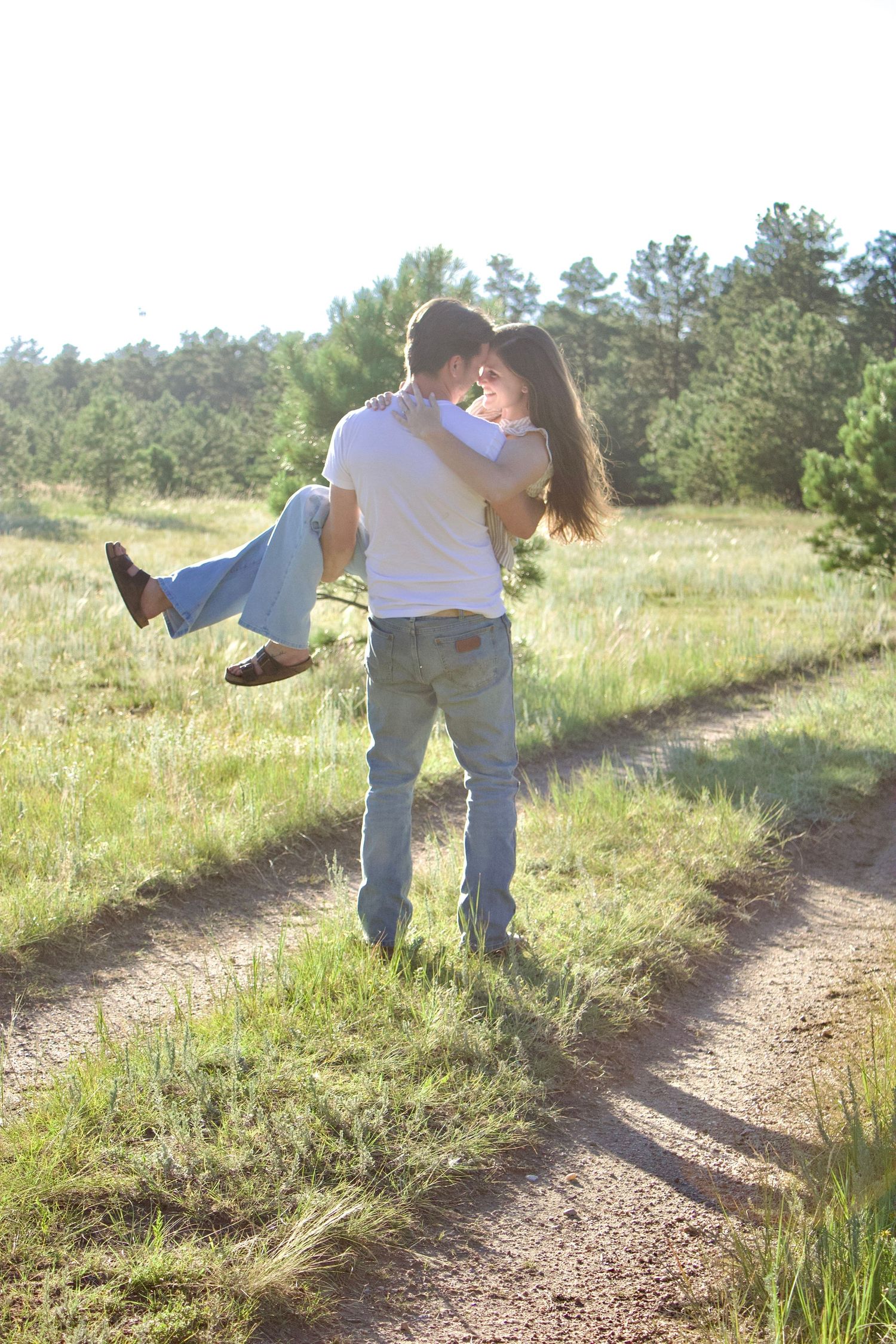 A couple in white clothing playfully dances together on a dirt path surrounded by green grass and trees.