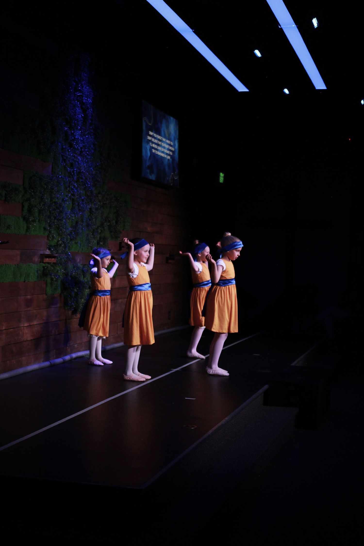 Young girls in orange dresses and blue headbands perform a dance routine on a dimly lit stage.