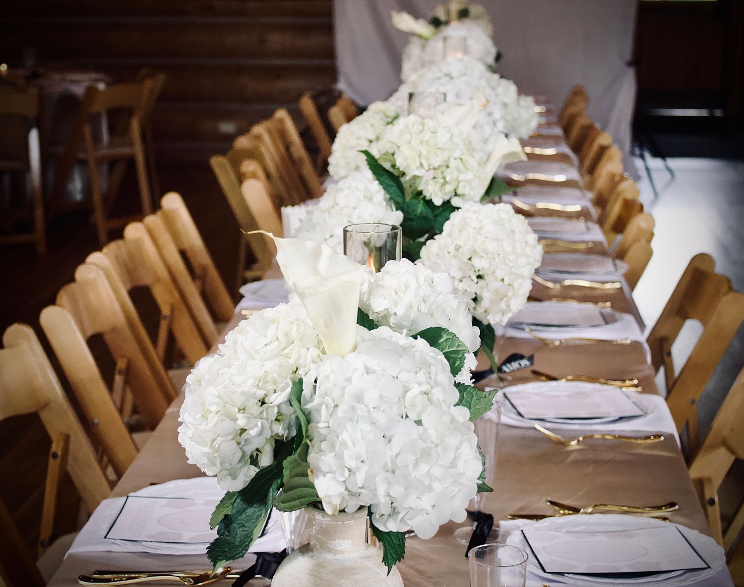 A long banquet table set for a wedding reception with white hydrangea centerpieces and wooden chairs.