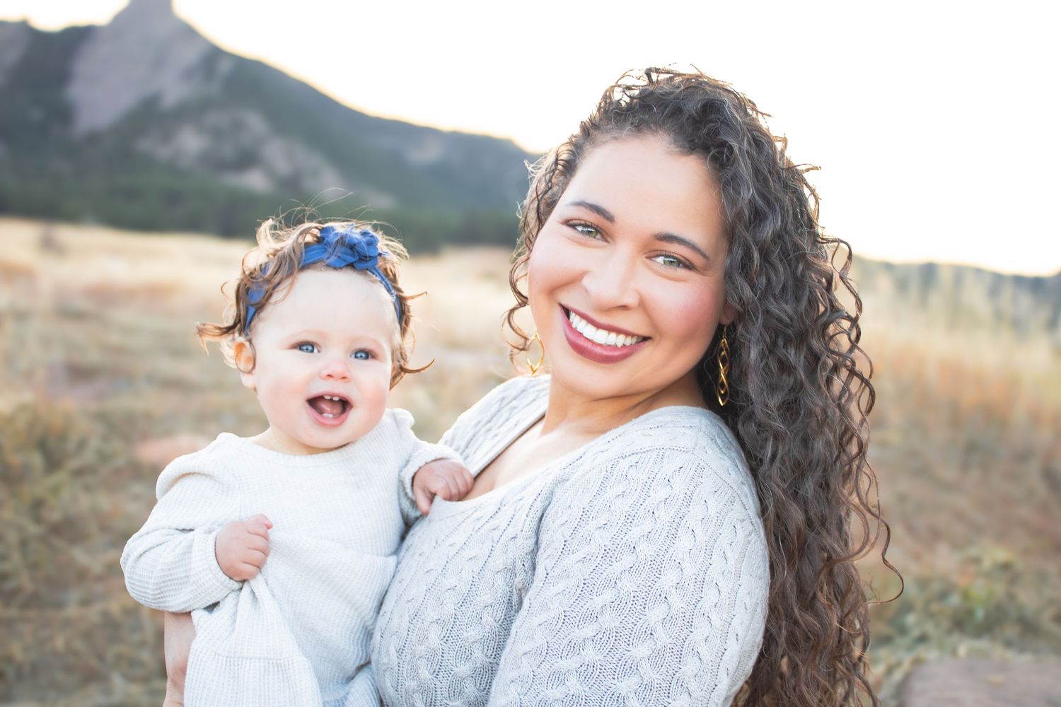 A joyful outdoor portrait of a parent and baby wearing light colored clothing with mountains in the background.