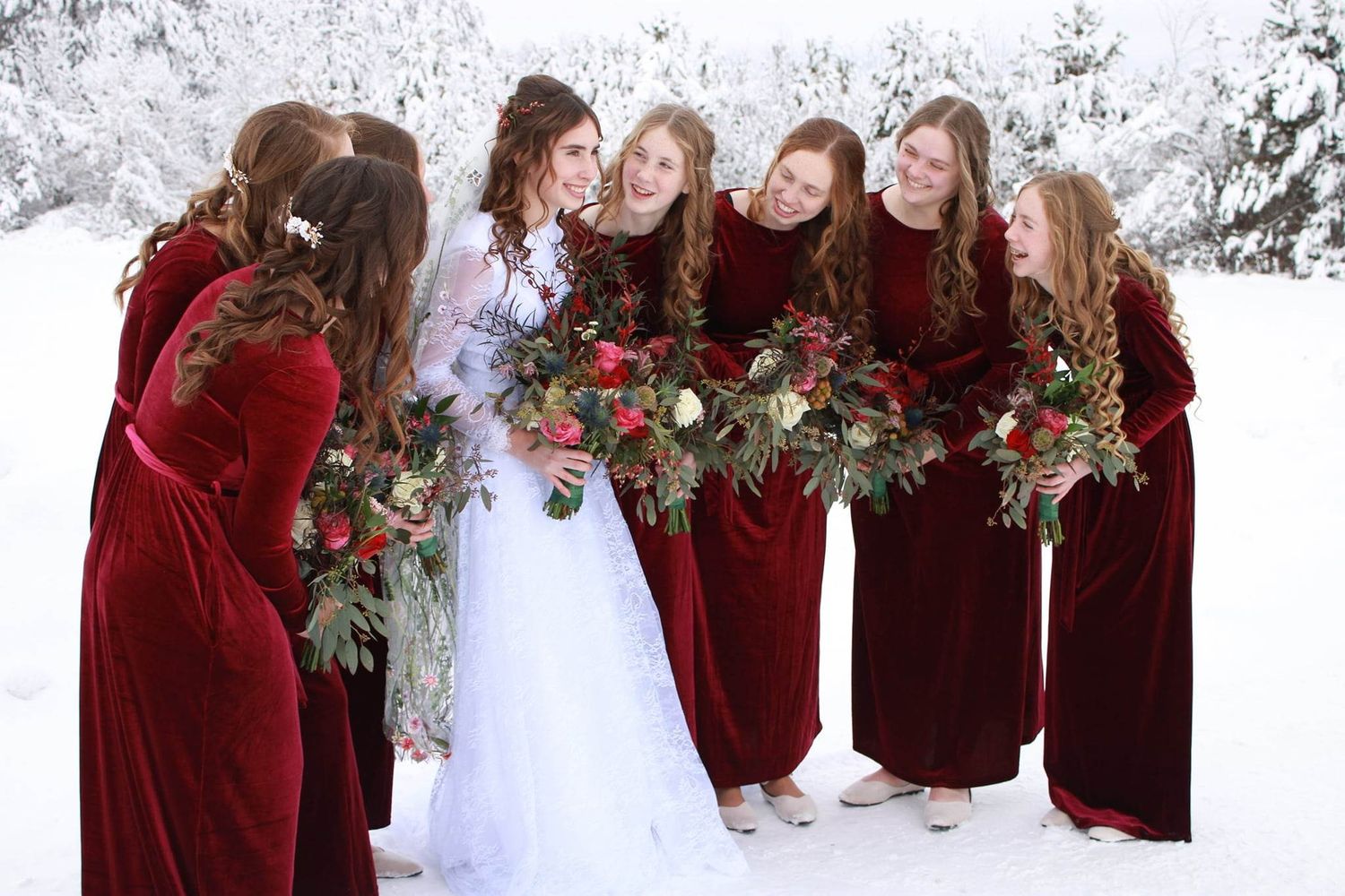 Bridesmaids in burgundy velvet dresses huddle together with bouquets in a snowy winter setting.