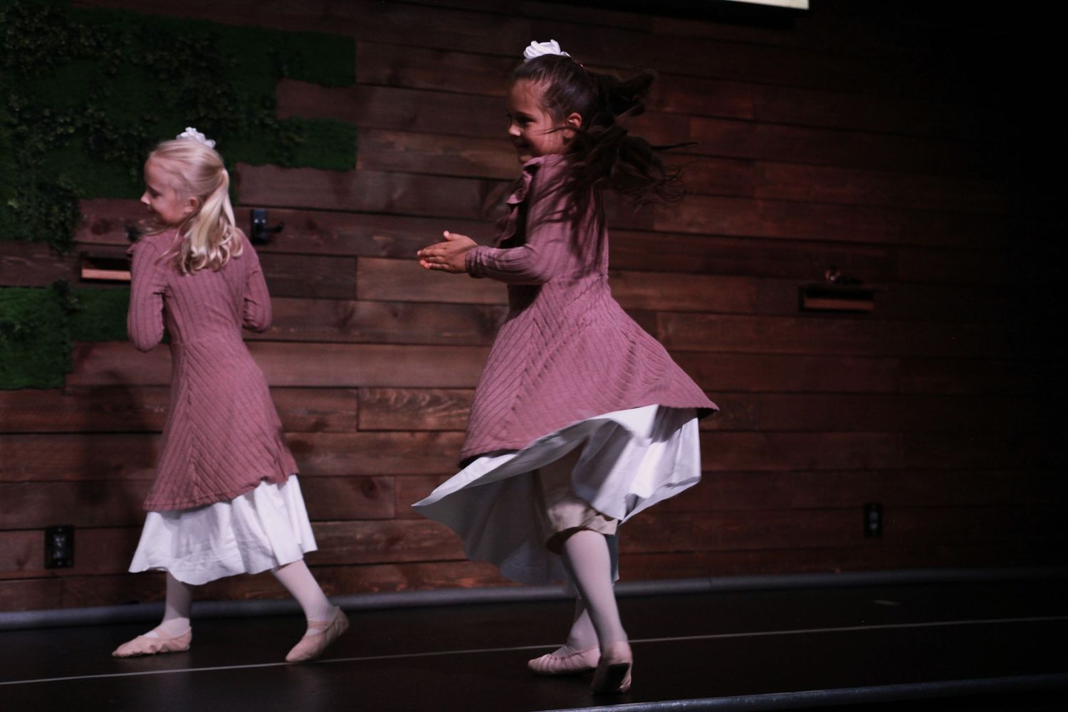 Two young girls in vintage pink dresses perform a dance routine on a dimly lit stage.