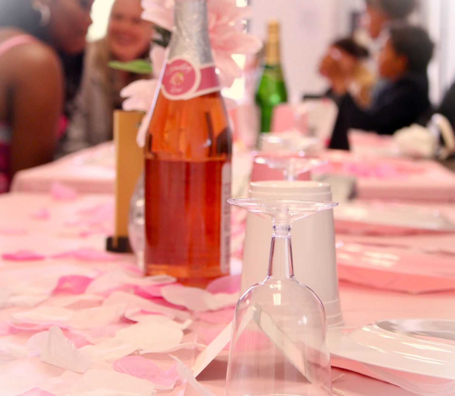 Pink-tinted party table setting with champagne bottles and glasses on a rose-petal covered surface.