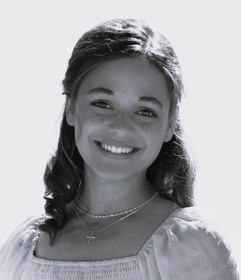 Smiling young woman with dark hair wearing a cross necklace and white top in a black and white portrait photo.