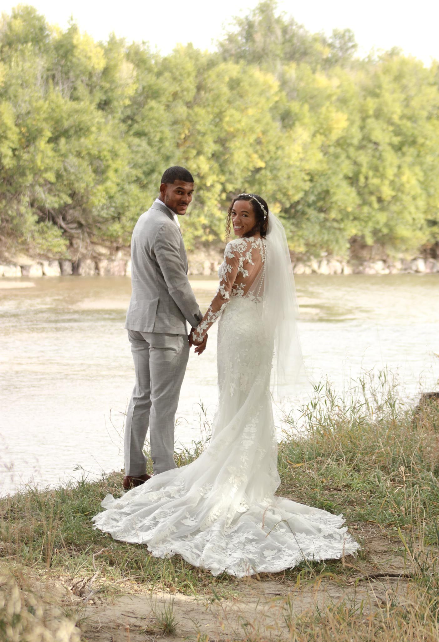 A couple in wedding attire shares a romantic moment by a riverside in soft natural lighting.