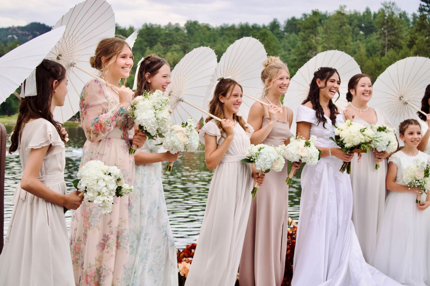 Wedding party holding white parasols stands in a row against a scenic mountain lake backdrop.