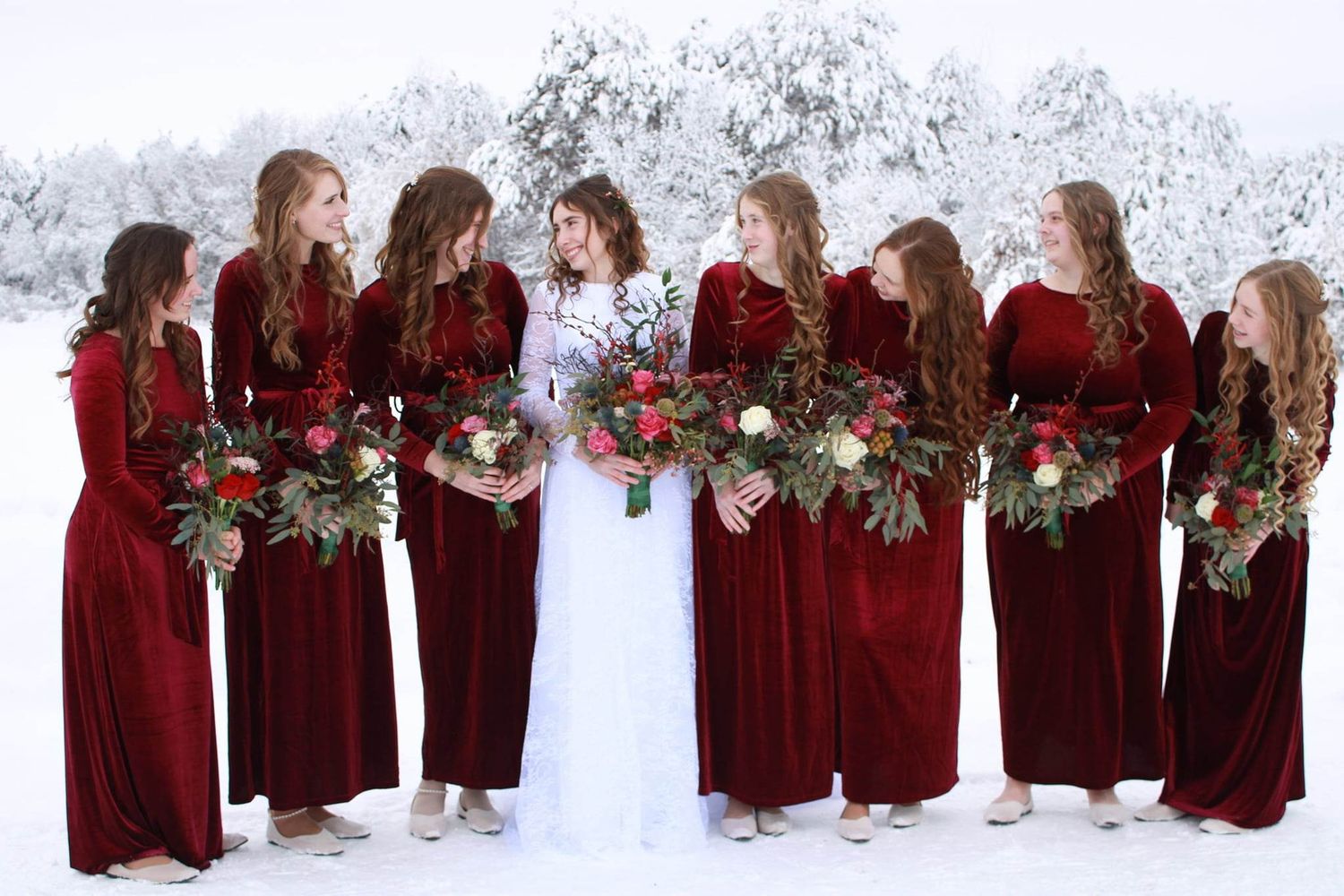 Wedding party in burgundy velvet and white posing together against snowy winter backdrop.