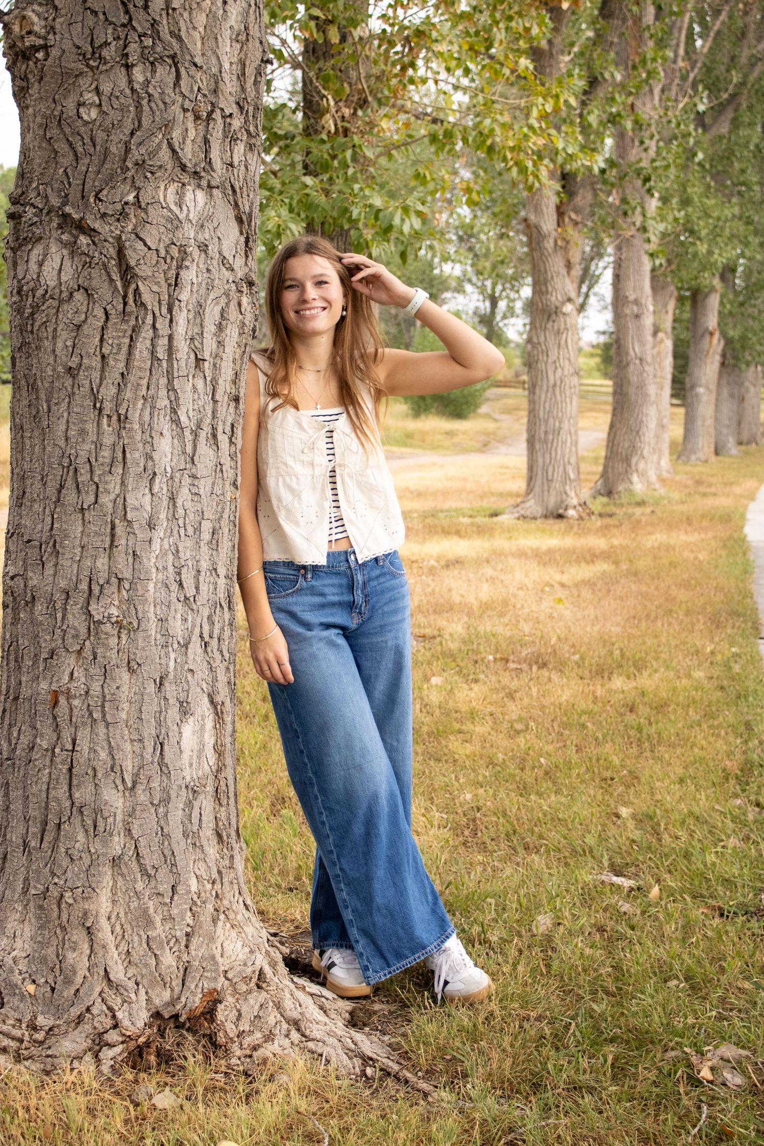 A person in jeans and striped top poses playfully behind a row of large tree trunks in a sunny outdoor setting.