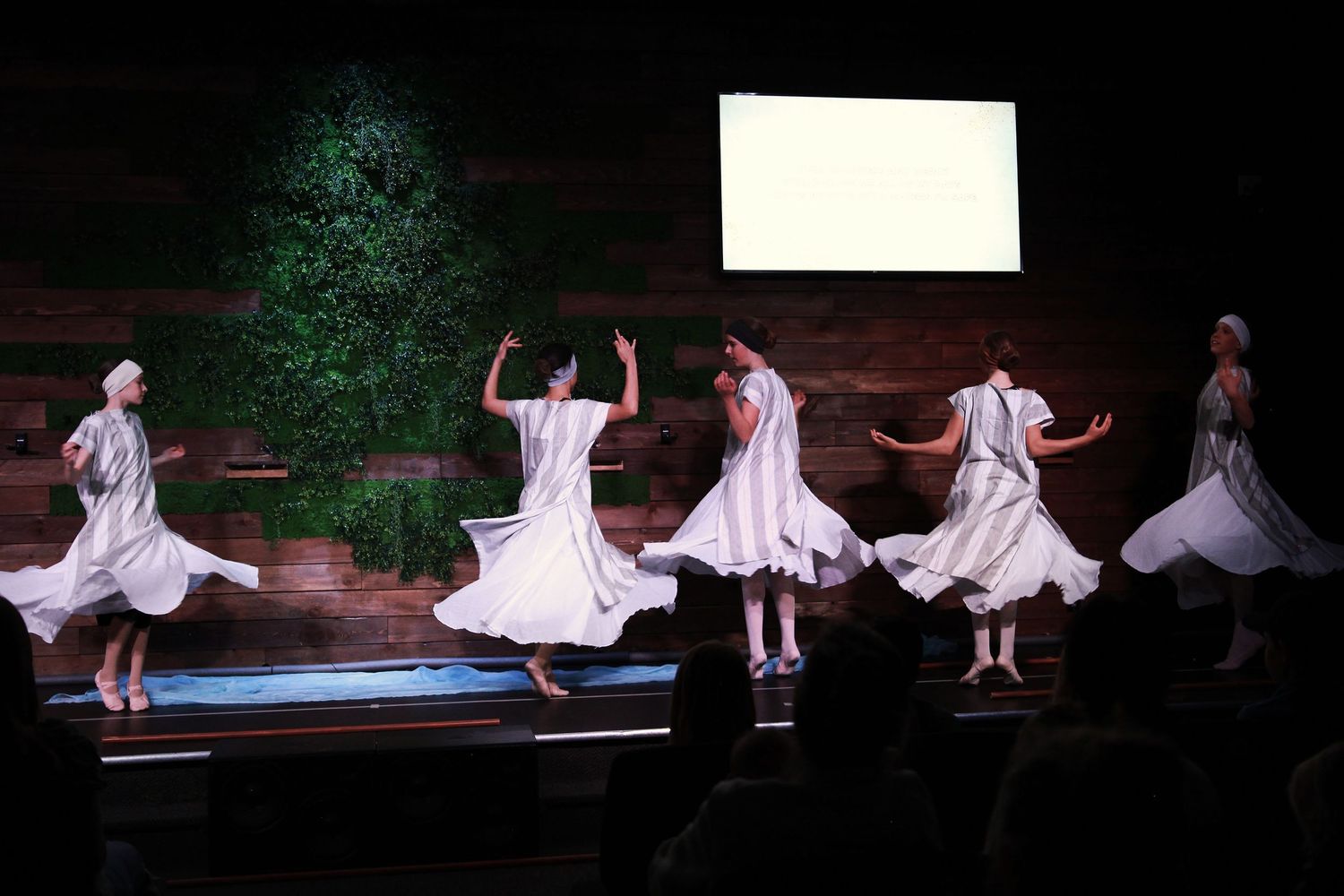 Women in white dresses perform a graceful whirling dance on a stage with green backdrop and screen.