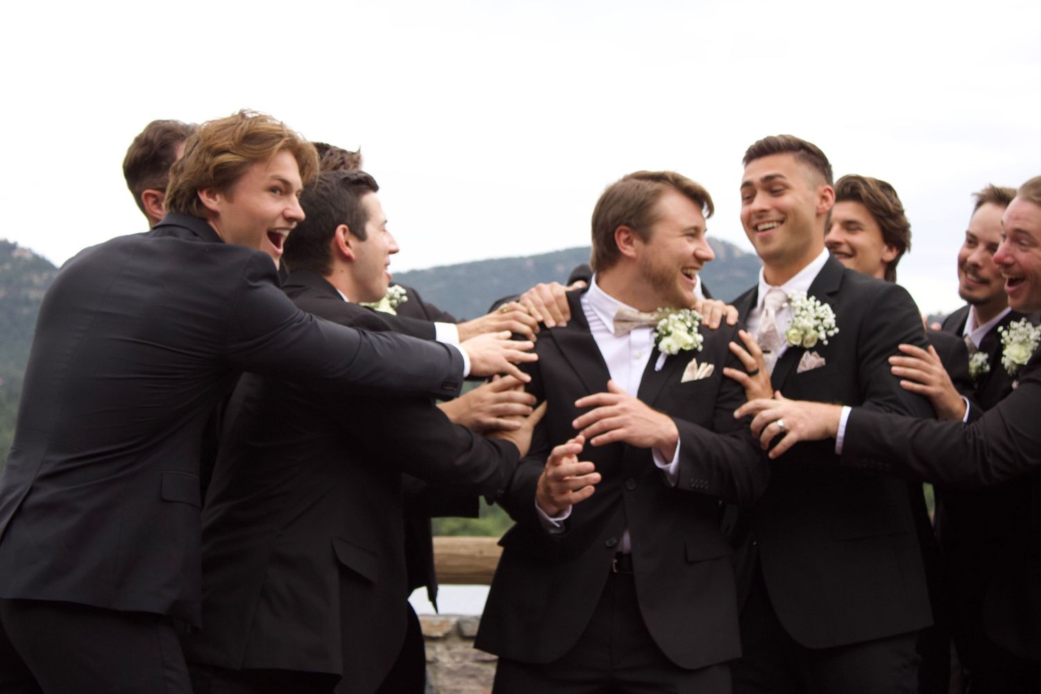 Wedding party members in black suits laugh and embrace during outdoor group photos at celebration.