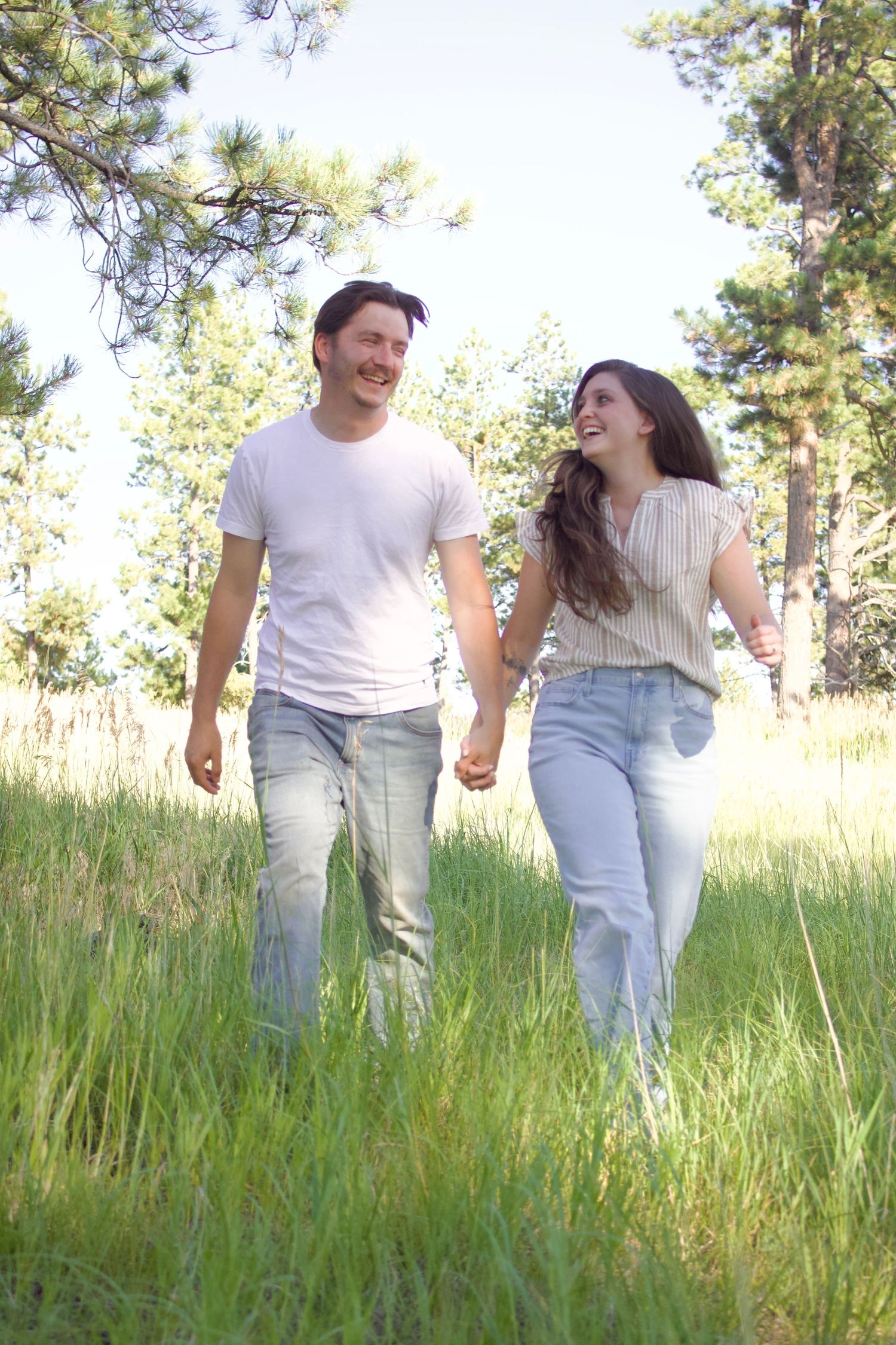 A couple runs joyfully through a sunny meadow with tall grass and pine trees in the background.