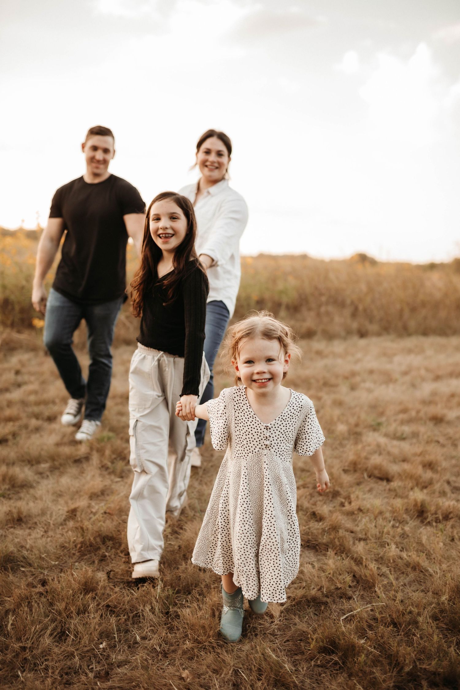 A family walks together through a golden field at sunset while their child runs ahead in a polka dot dress.