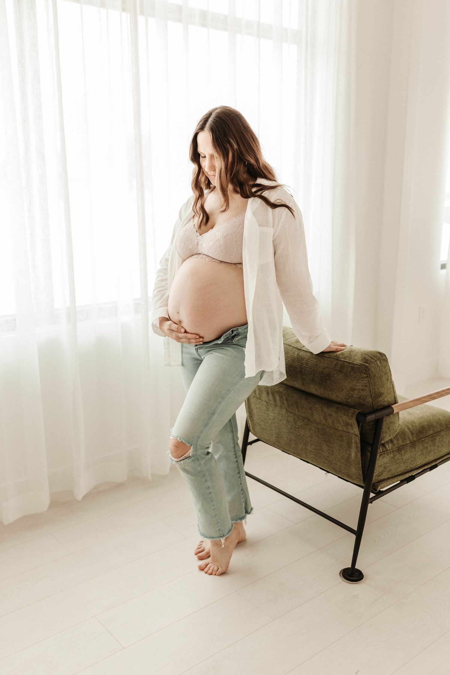 A person in a cream sweater and sage green pants sits casually on a modern olive velvet chaise lounge near white curtains.