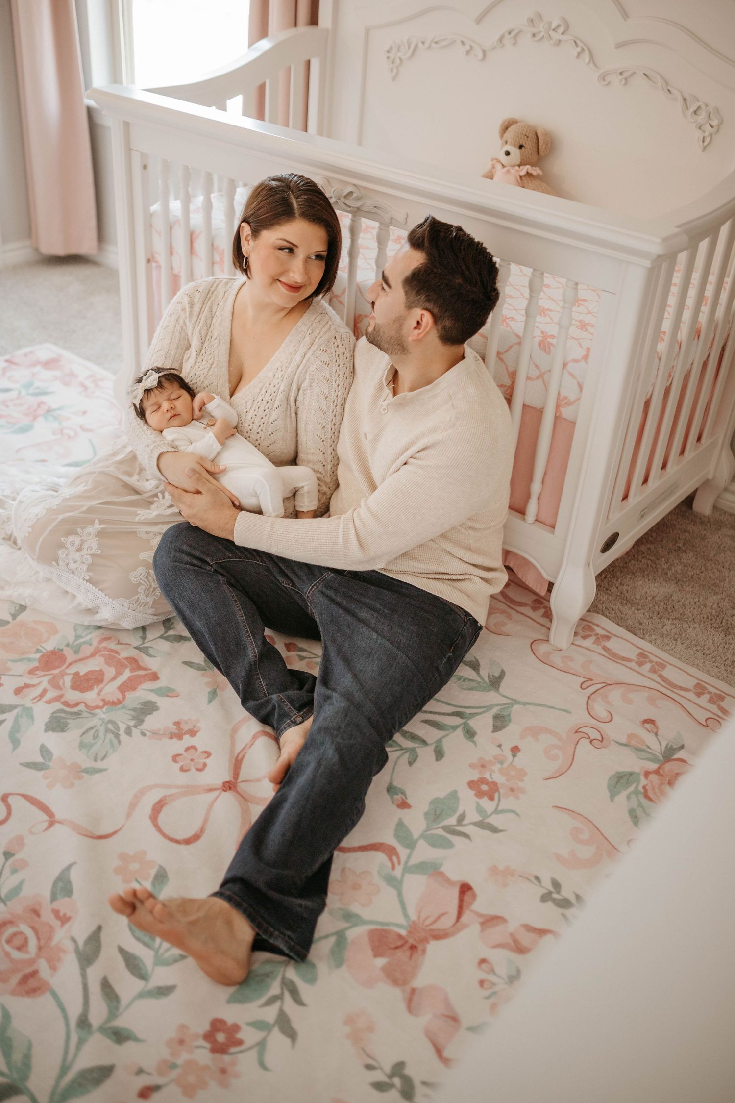Parents sit together on a floral rug in a nursery with soft pink walls and white furniture.