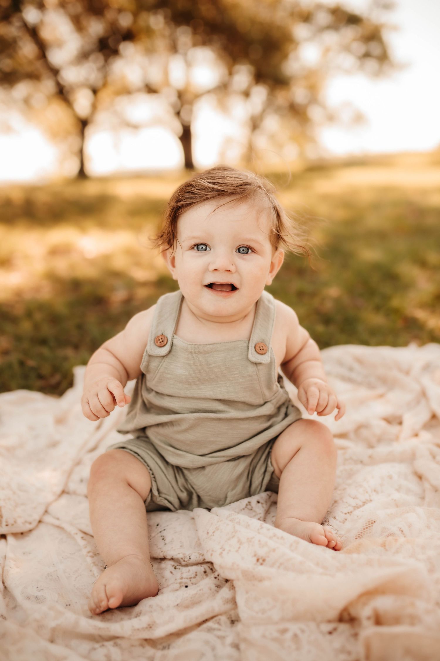 A baby wearing tan overalls sits on a blanket outdoors during golden hour with trees in the background.